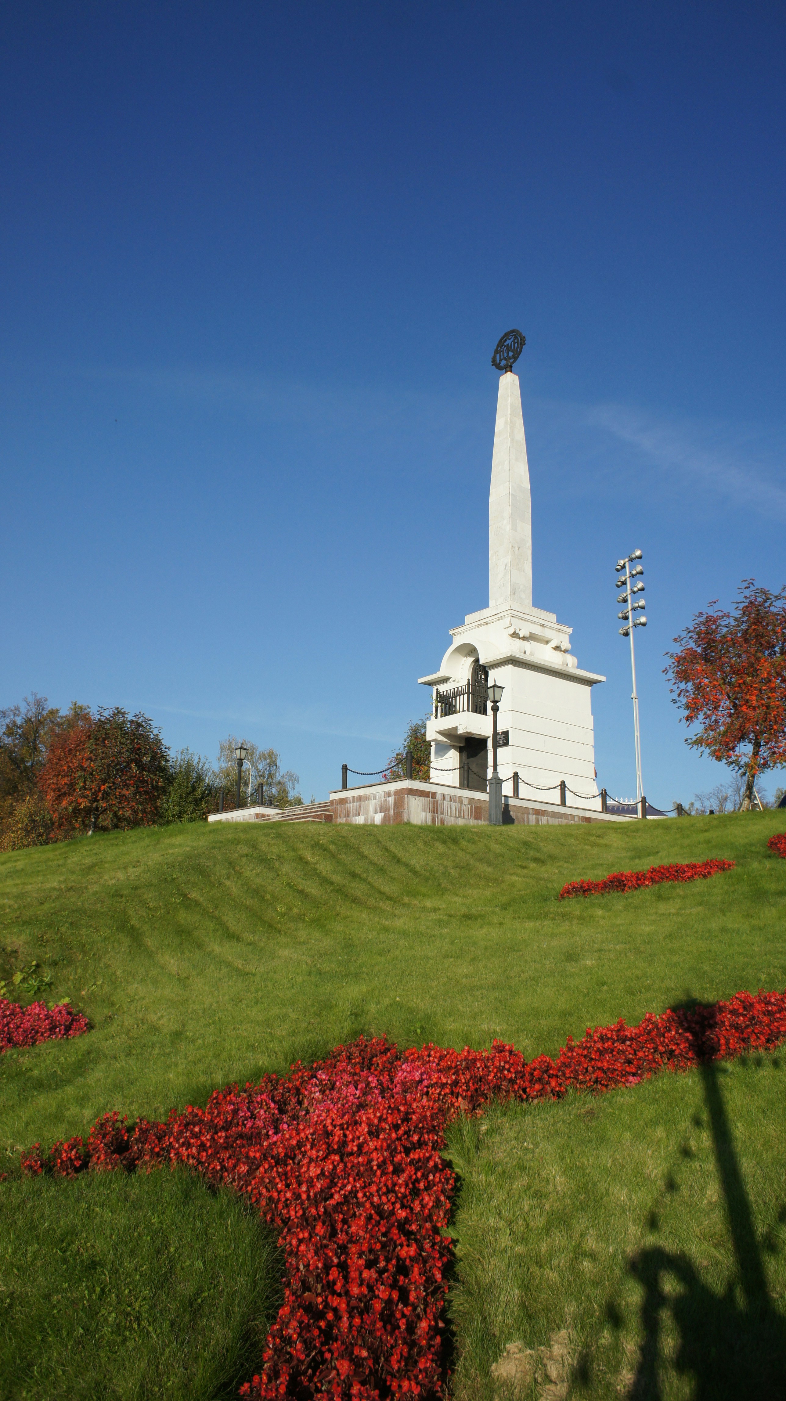 A white stone obelisk on a grassy hill with red flower beds