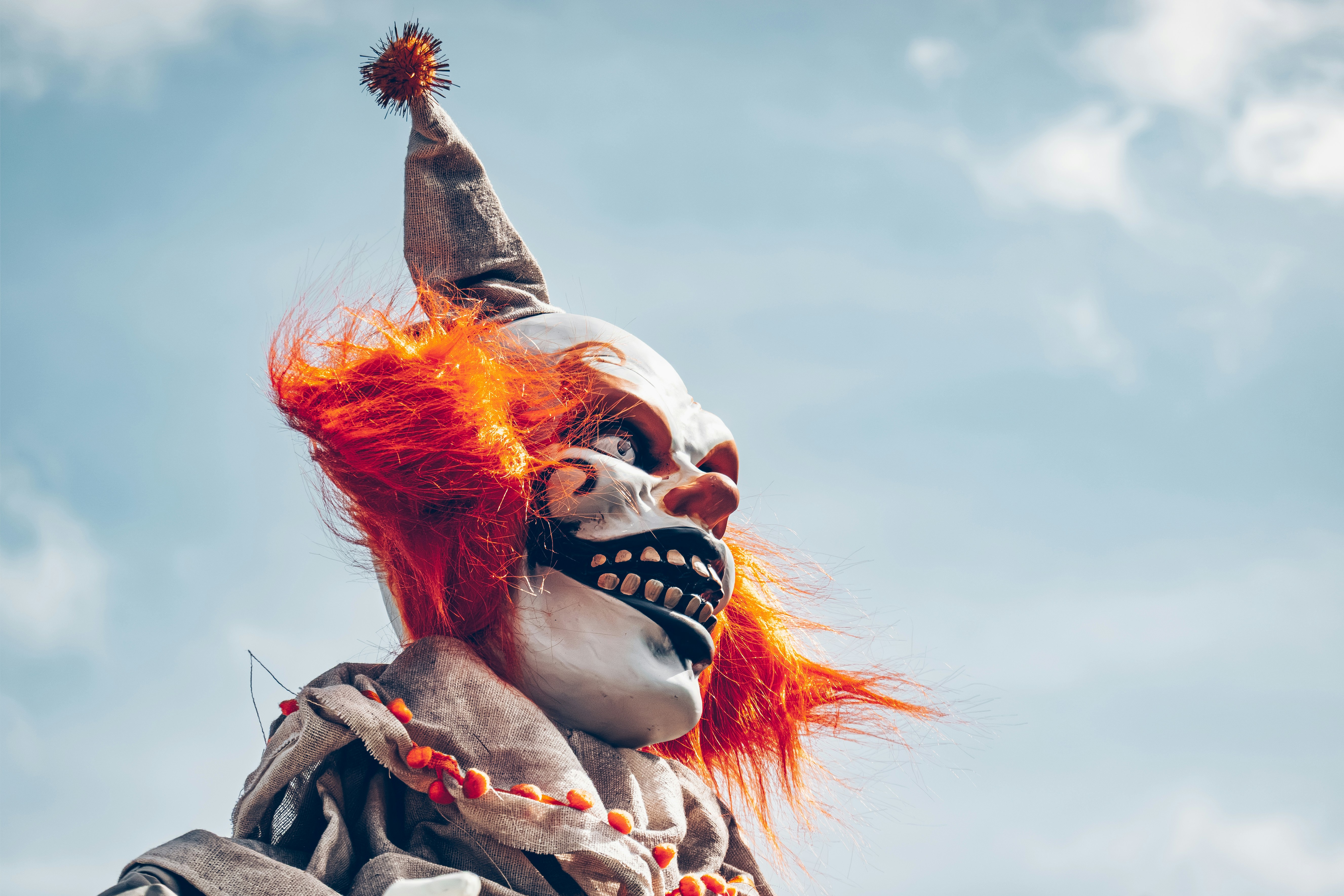 A clown mask with bright orange hair and a pointed hat against a blue sky