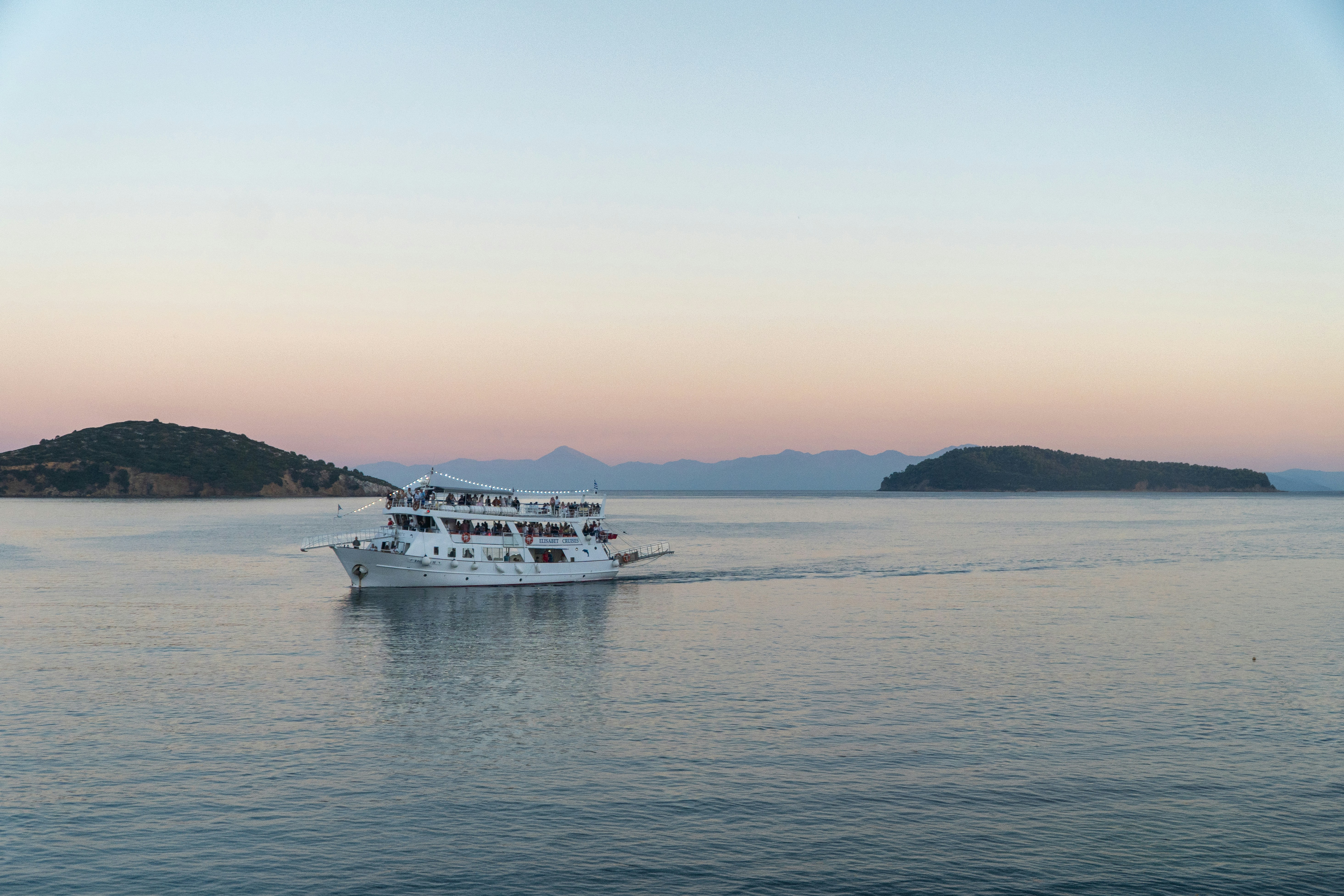 White tour boat crossing calm Aegean water at golden hour near island silhouettes