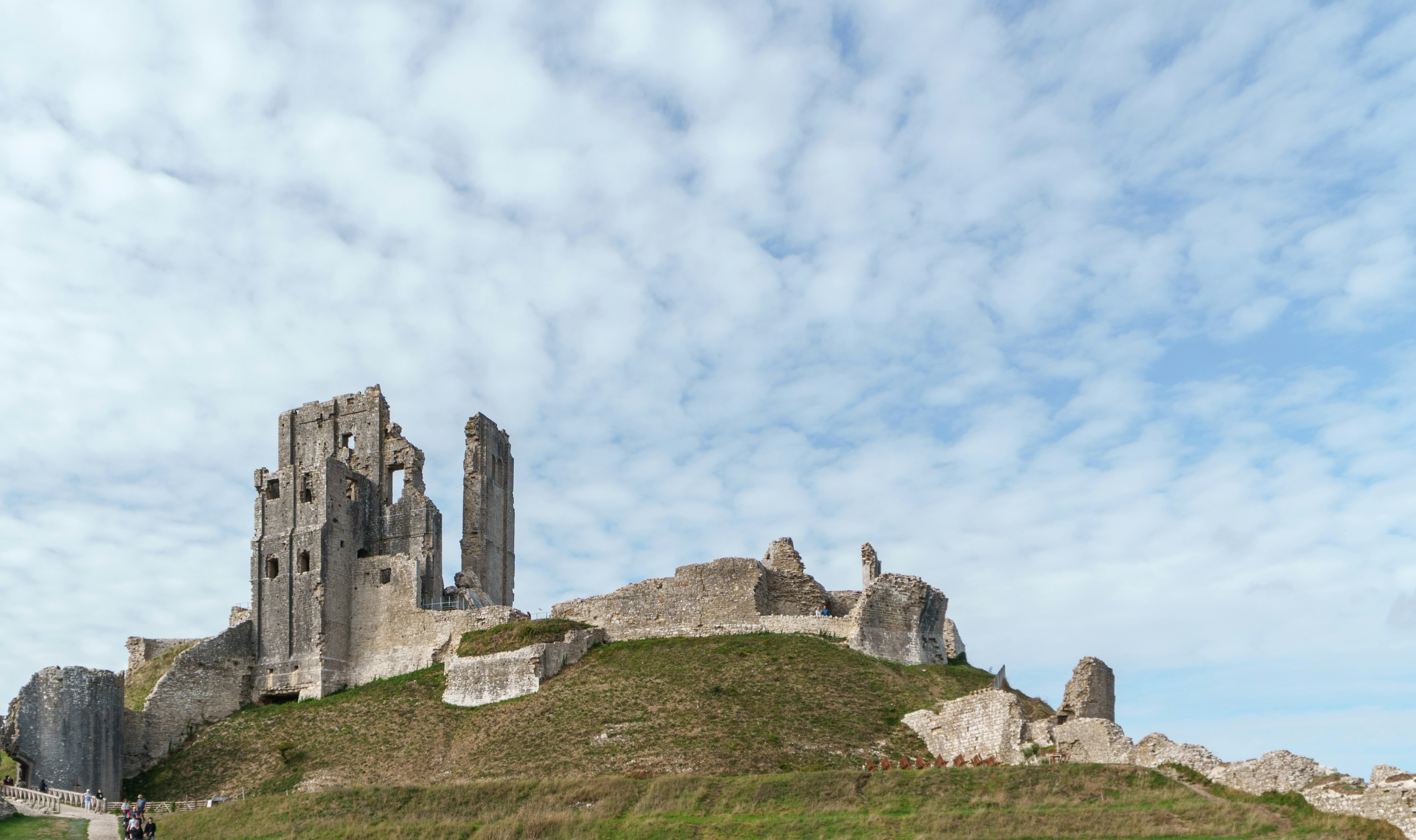 Stone ruins of Corfe Castle on a grassy hill under a cloudy blue sky