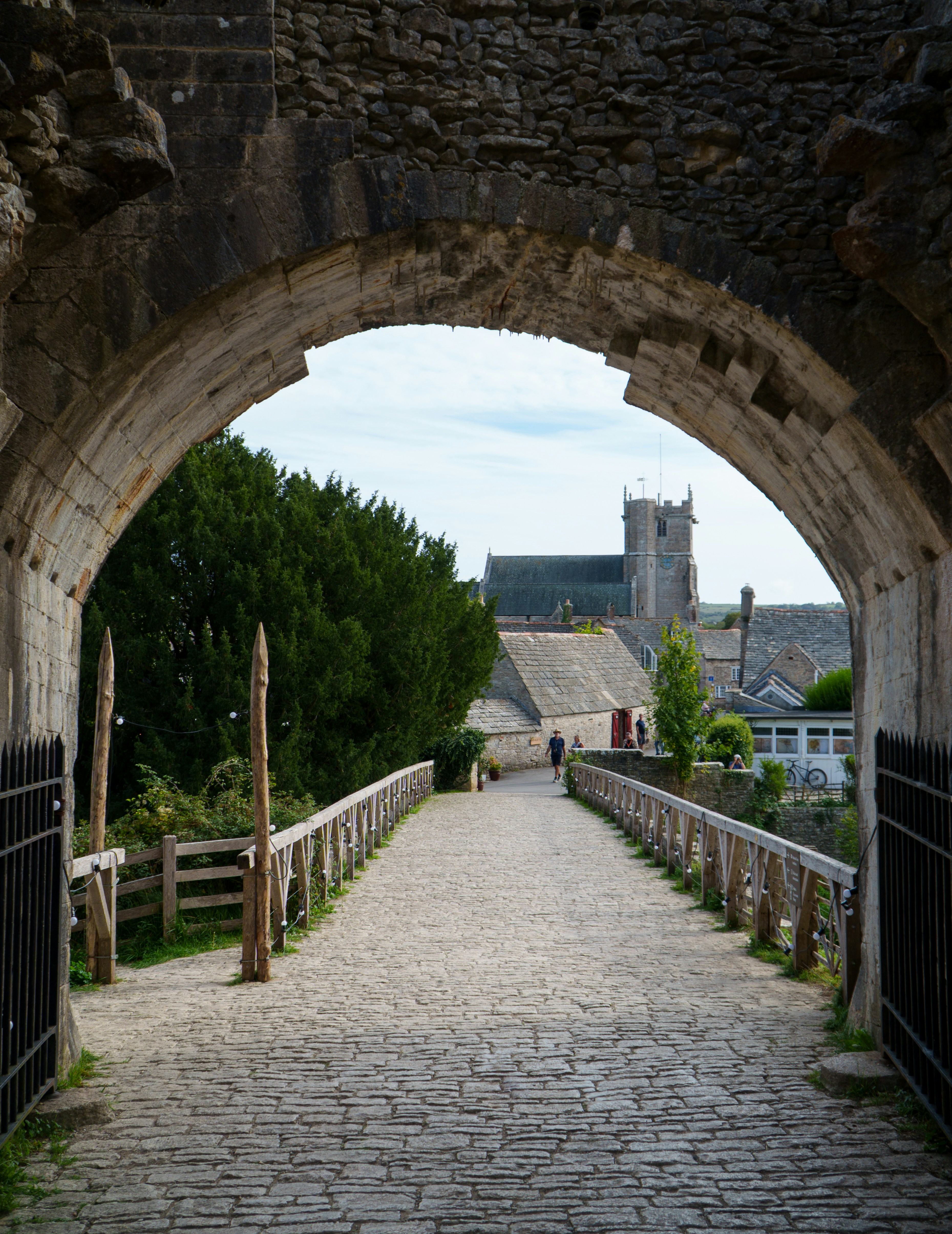 A stone bridge leading through an archway toward a village with a church tower