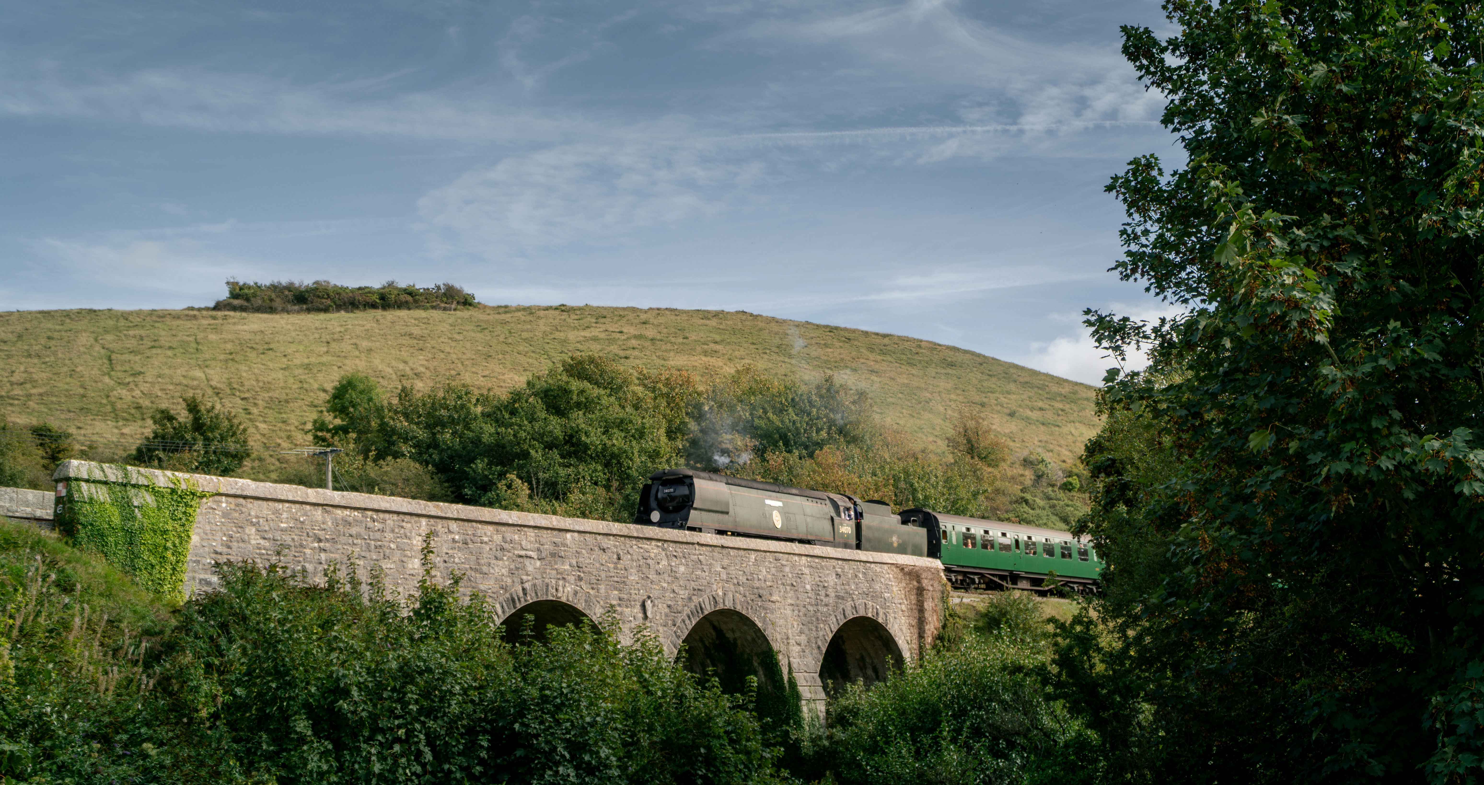 A steam train crossing a stone arch bridge below a grassy hill