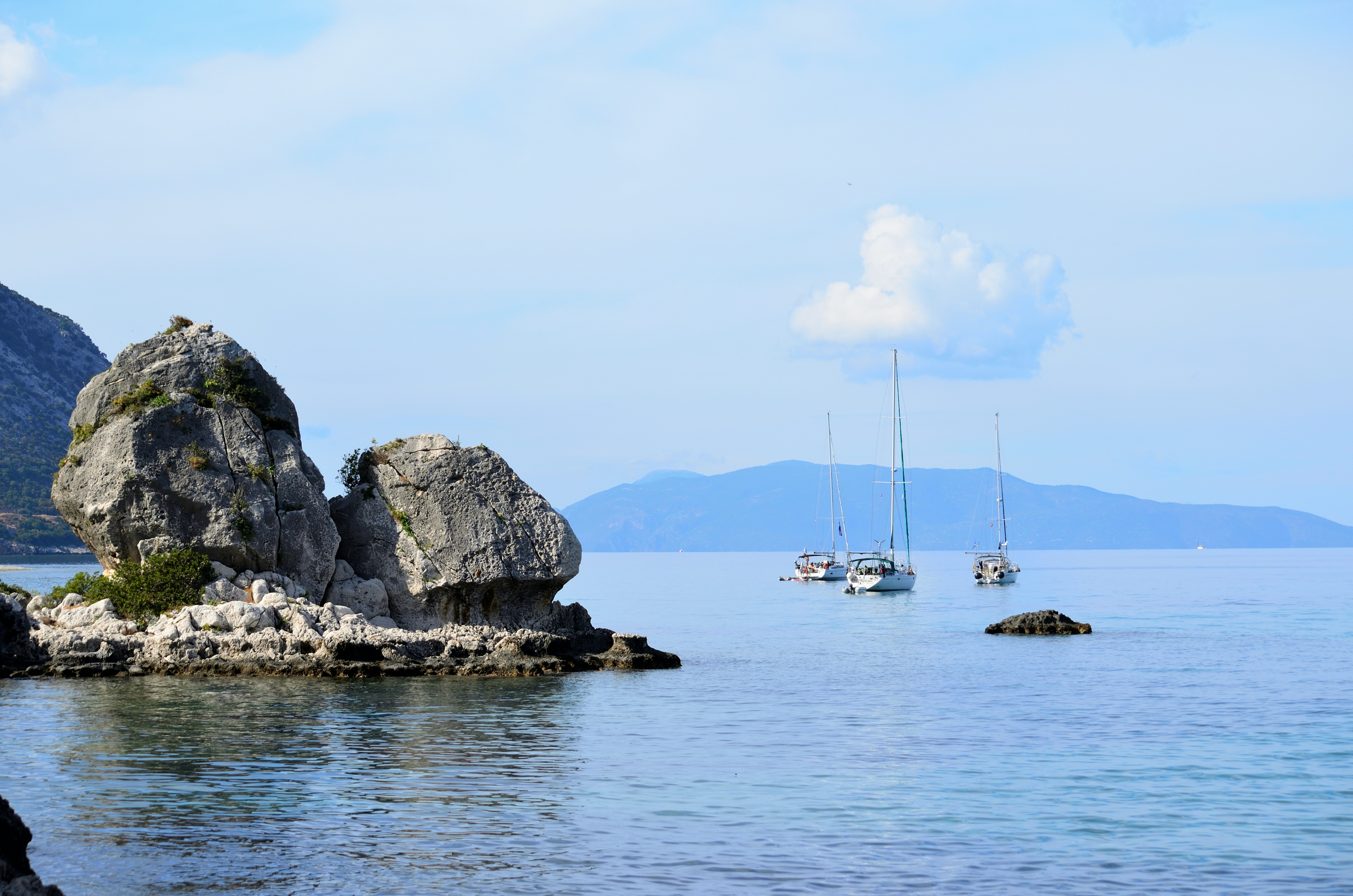Three sailboats on calm blue water near a large rocky outcrop