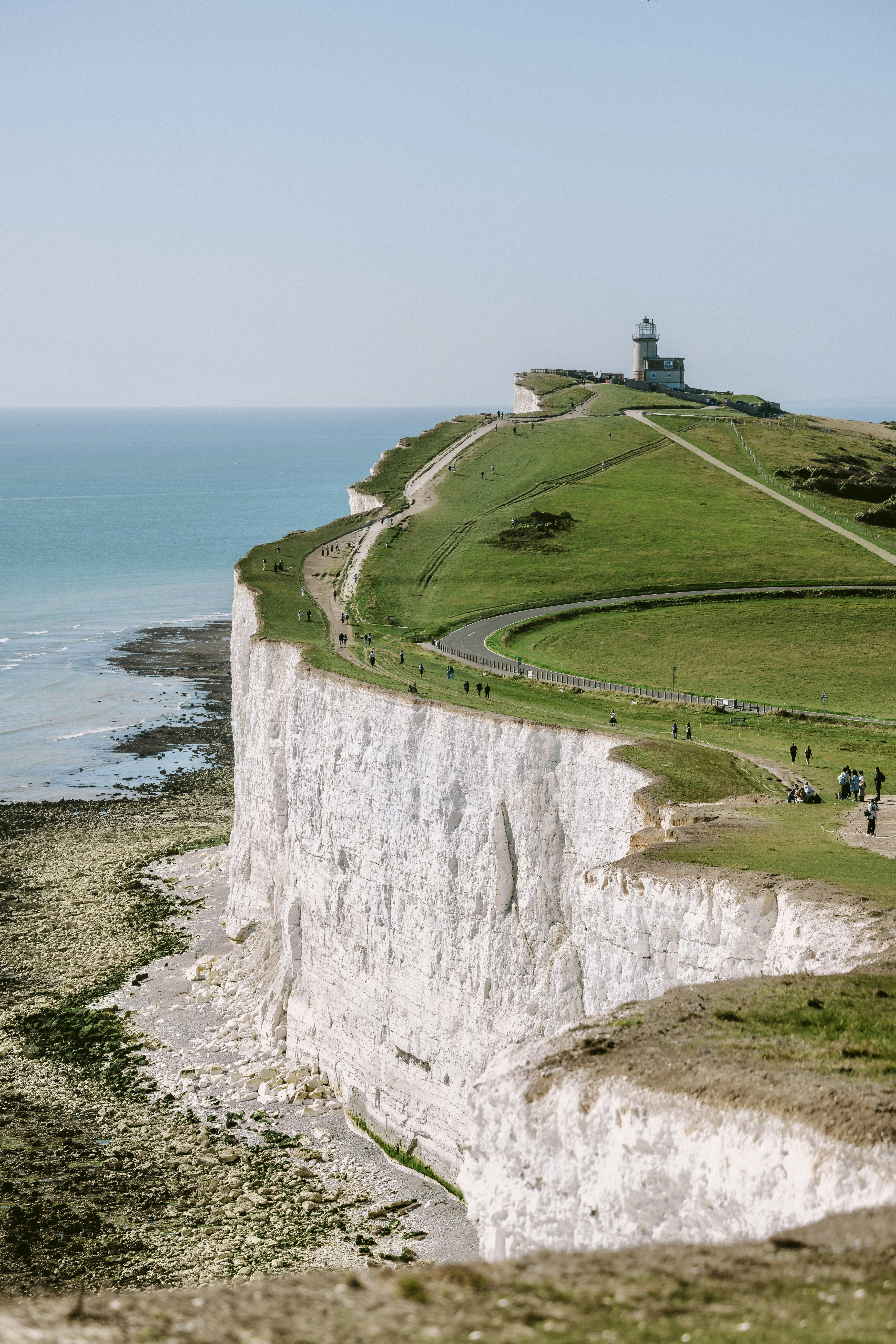 A lighthouse on white chalk cliffs above the sea with people walking nearby