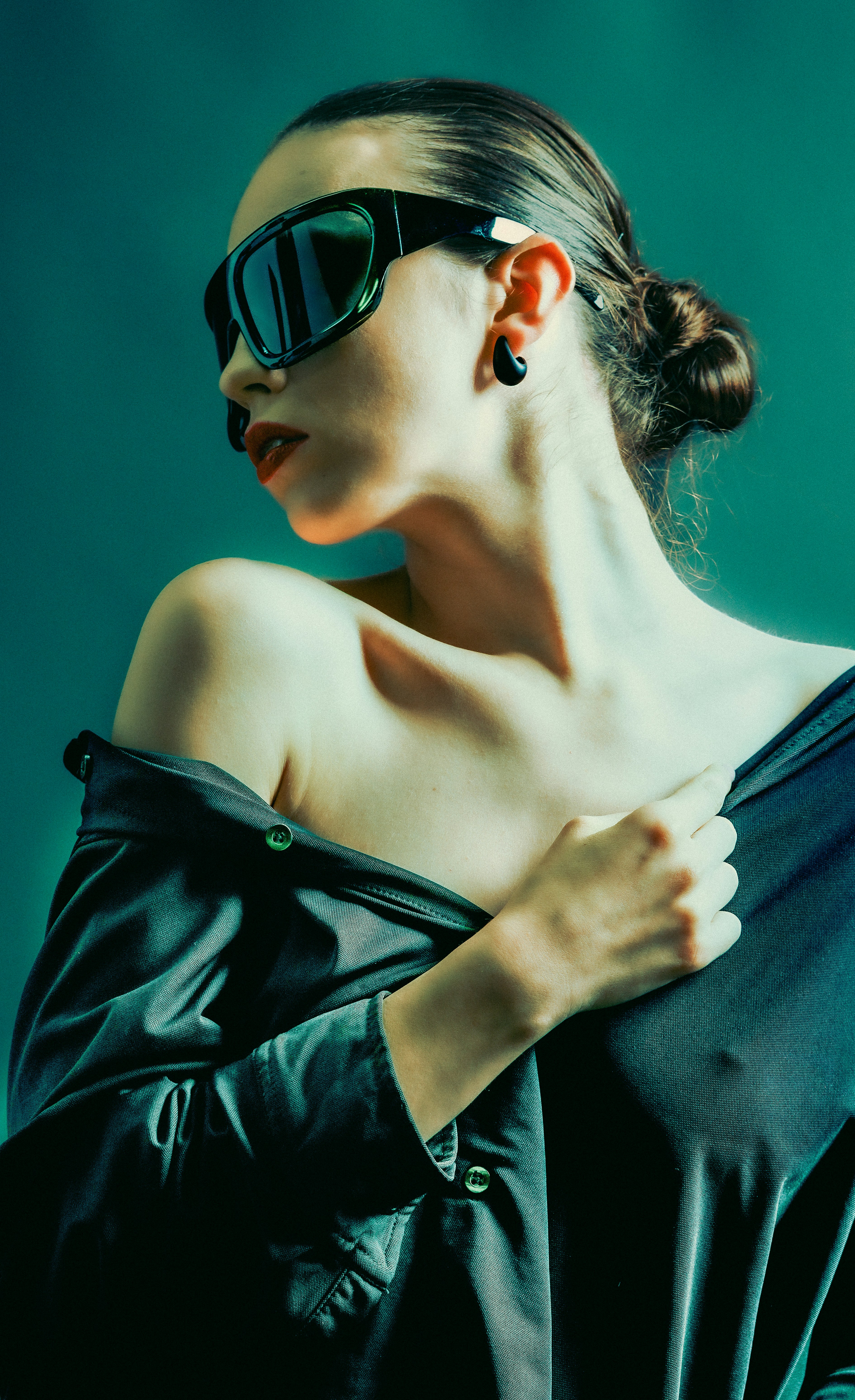 A woman in dark sunglasses and a buttoned shirt against a teal background