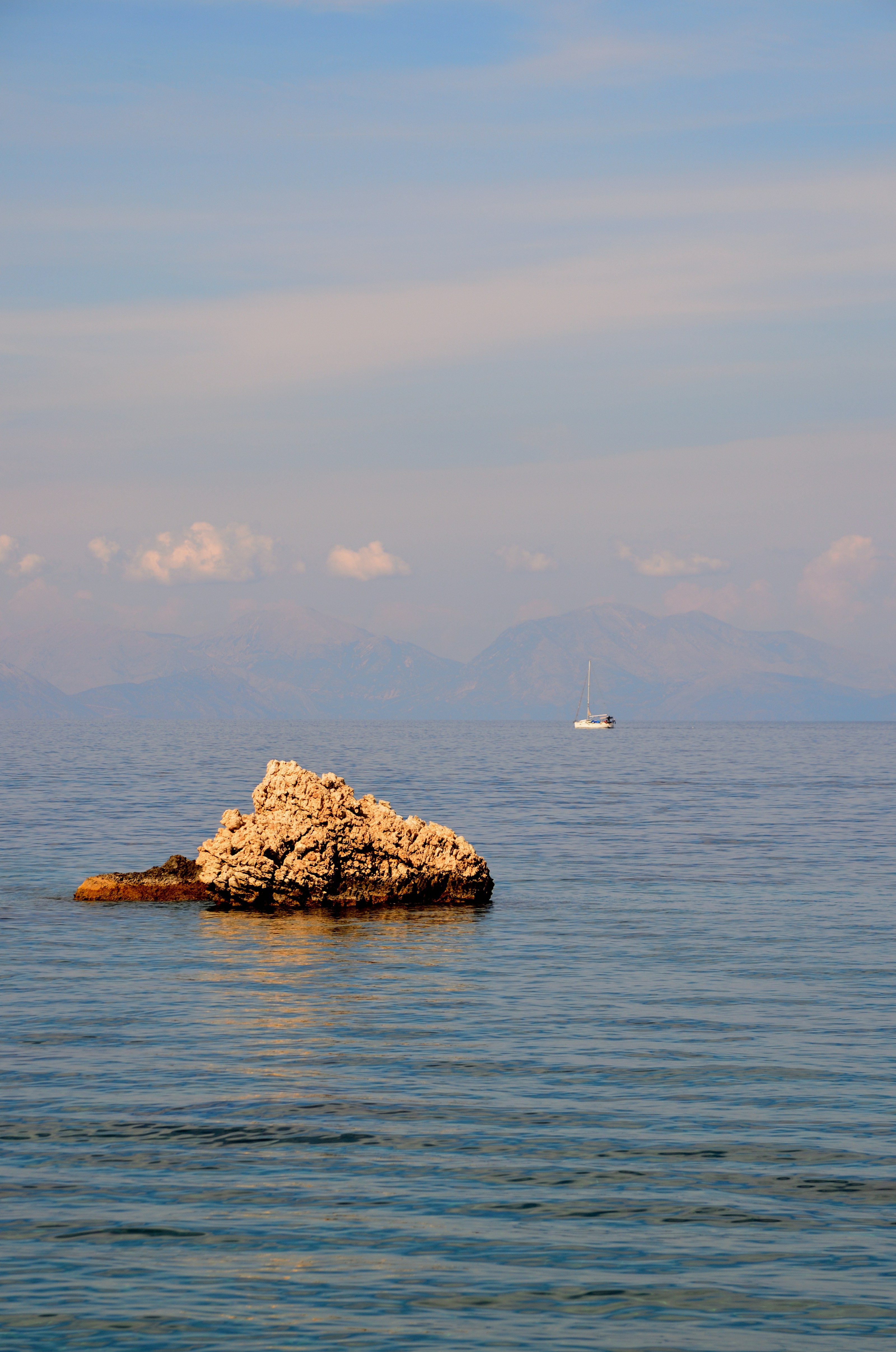 A jagged rock in calm blue water with a distant sailboat and mountains