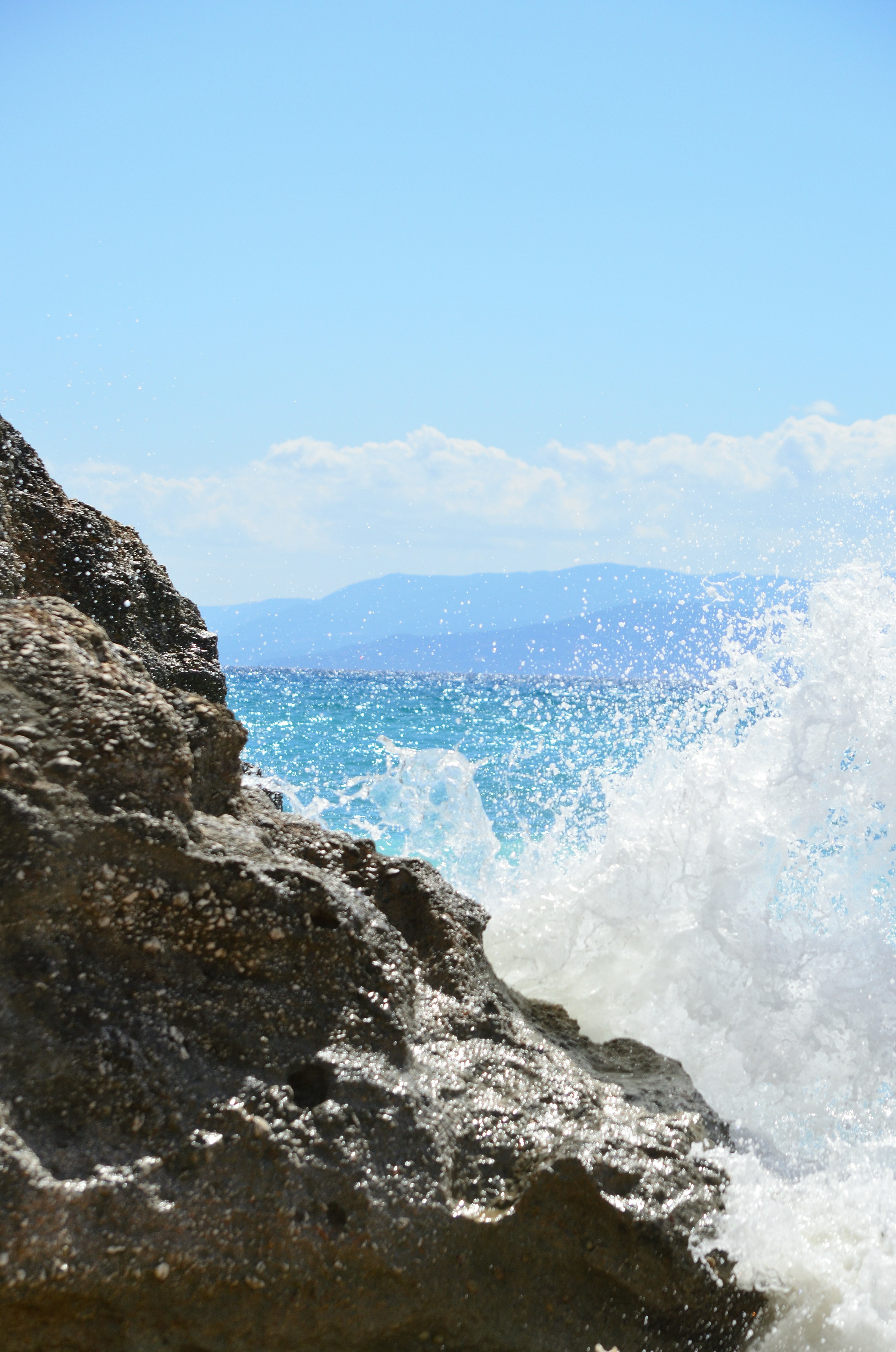 White sea spray crashing against a dark rocky cliff under a clear blue sky
