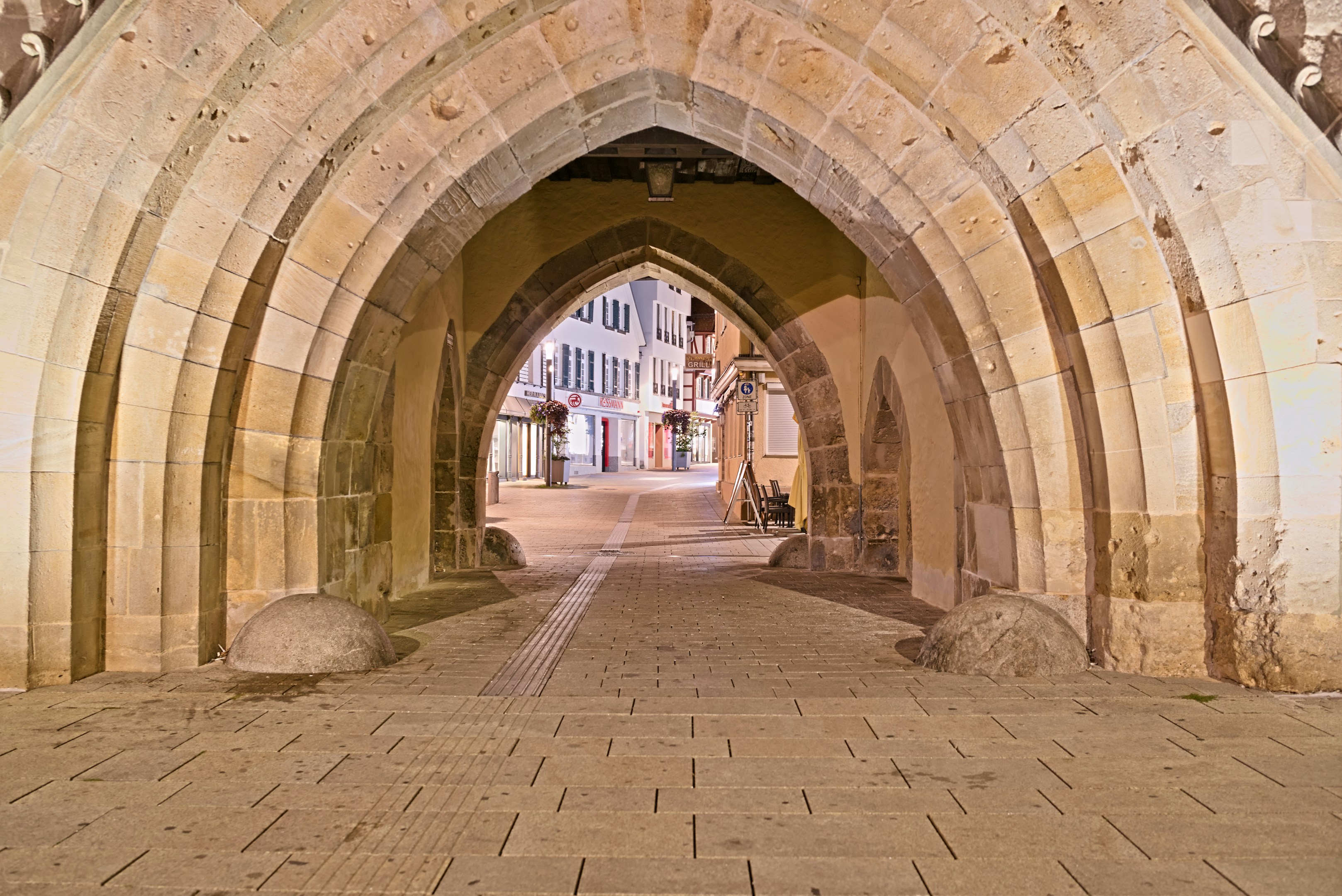 A stone archway leading to a quiet street with illuminated shopfronts at night