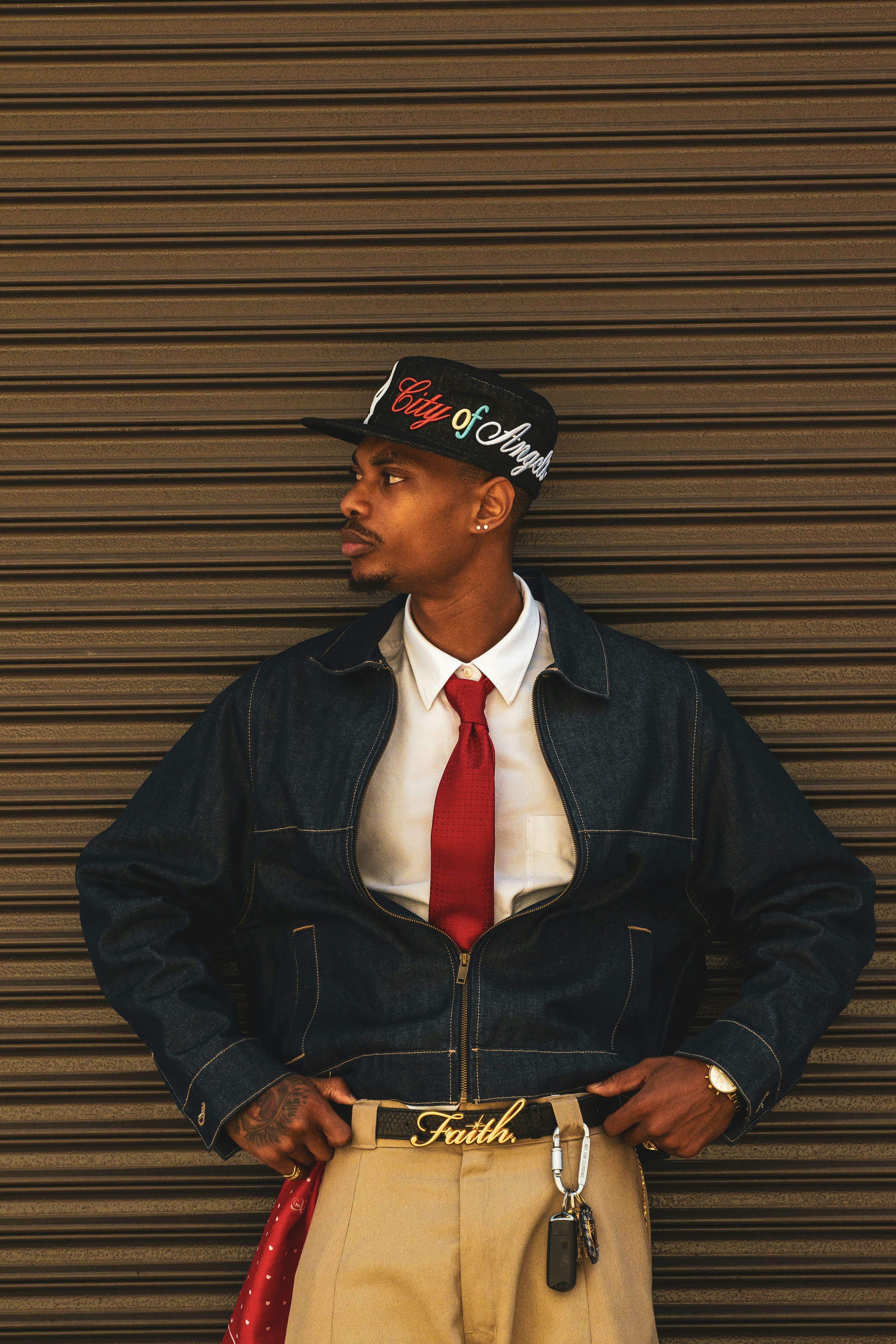 A man in a denim jacket, white shirt, red tie, and cap against a shutter