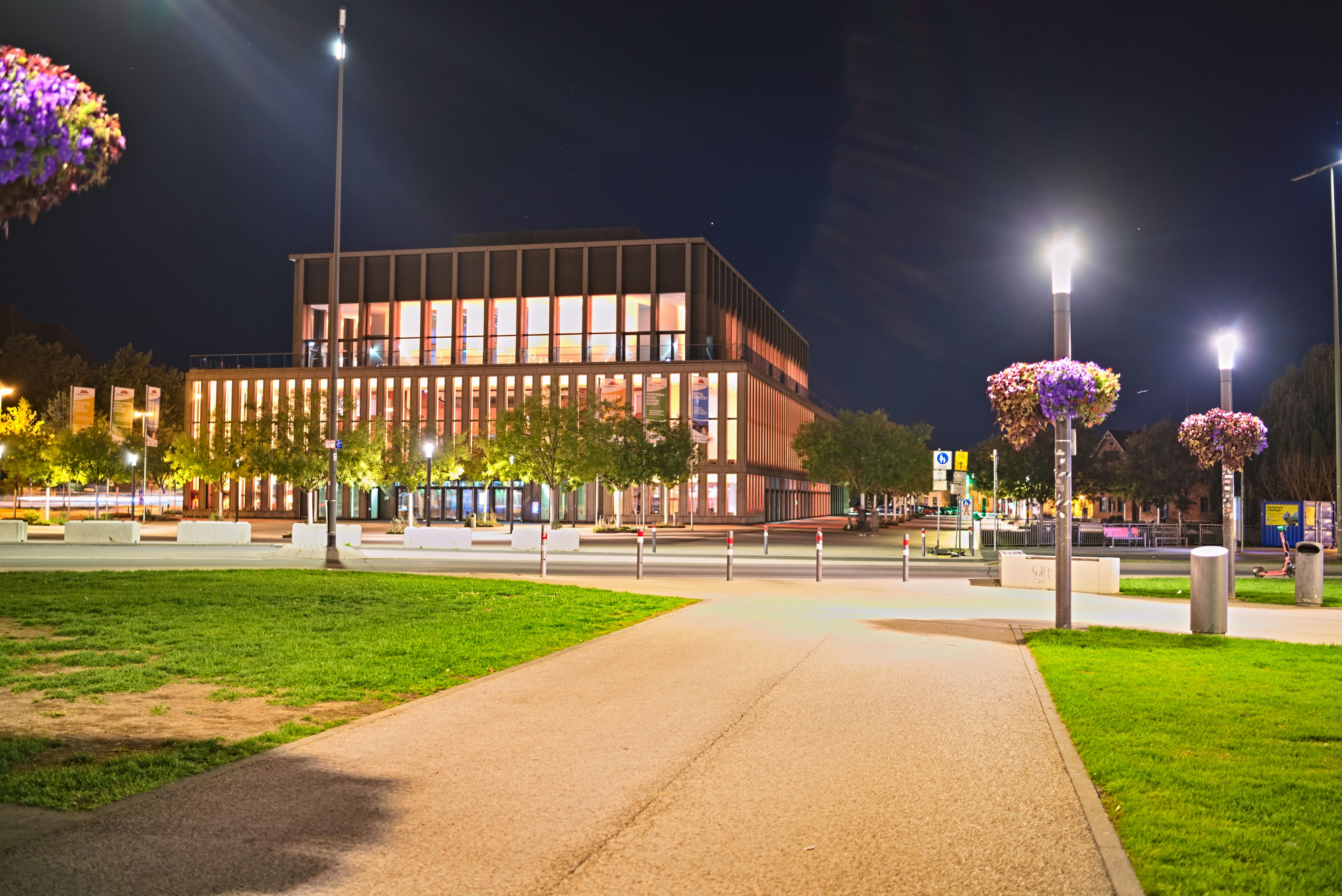 A modern building with illuminated windows at night beside a park with hanging flower baskets