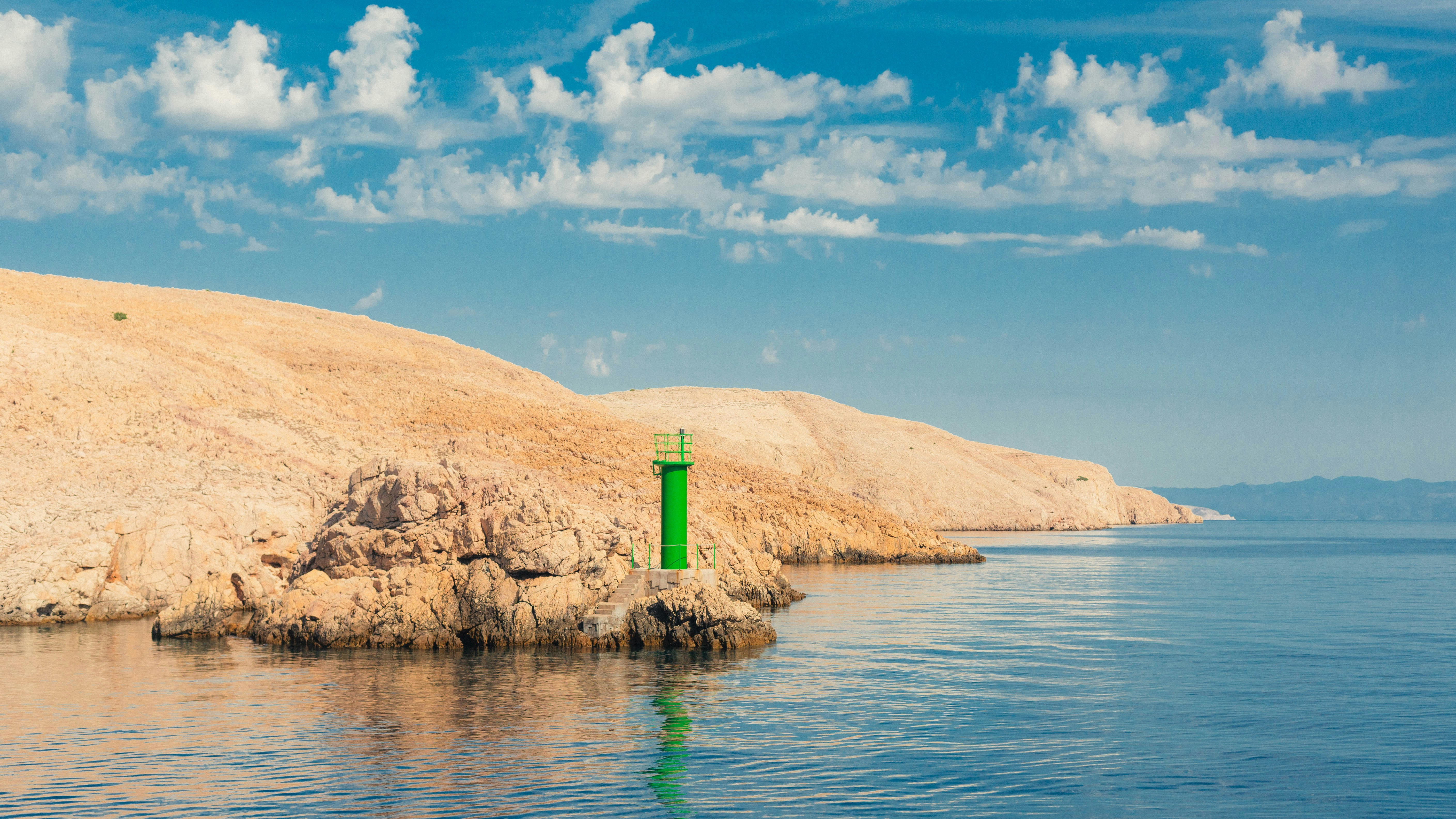 A green navigation beacon on a rocky coastline beside calm blue water