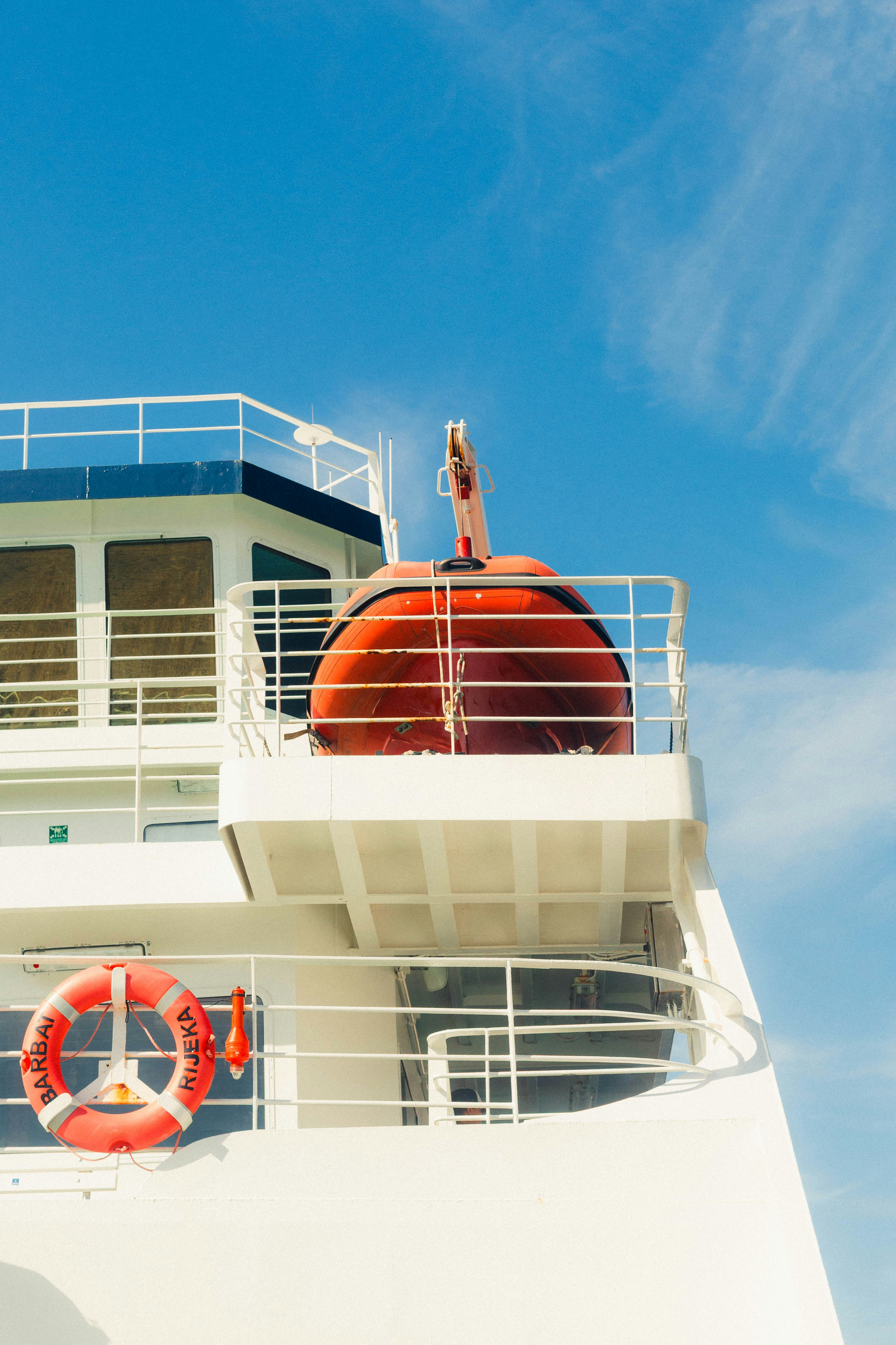 A white ferry deck with an orange life raft and lifebuoy against blue sky