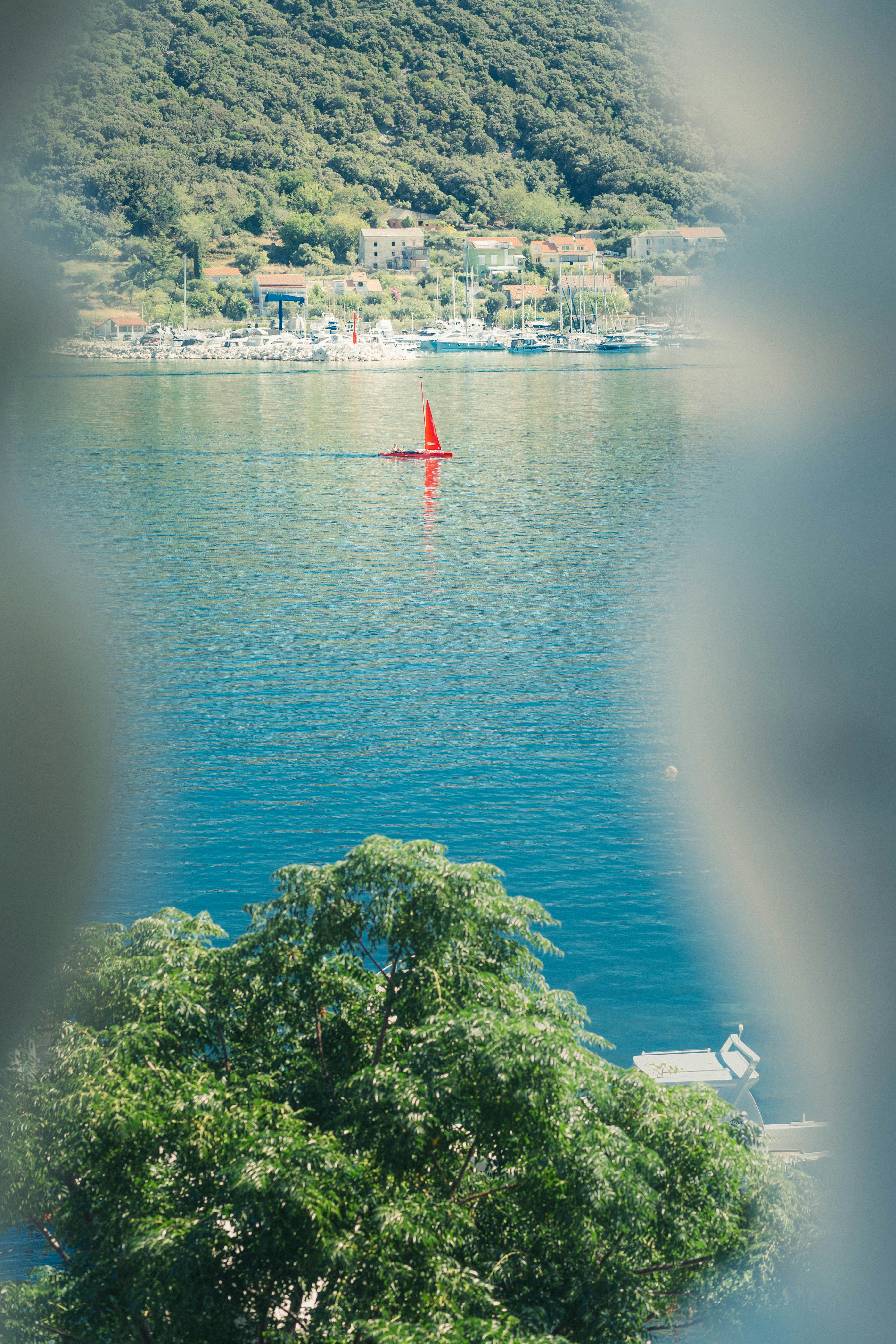 A small red sailboat on blue water near a coastal town and hills