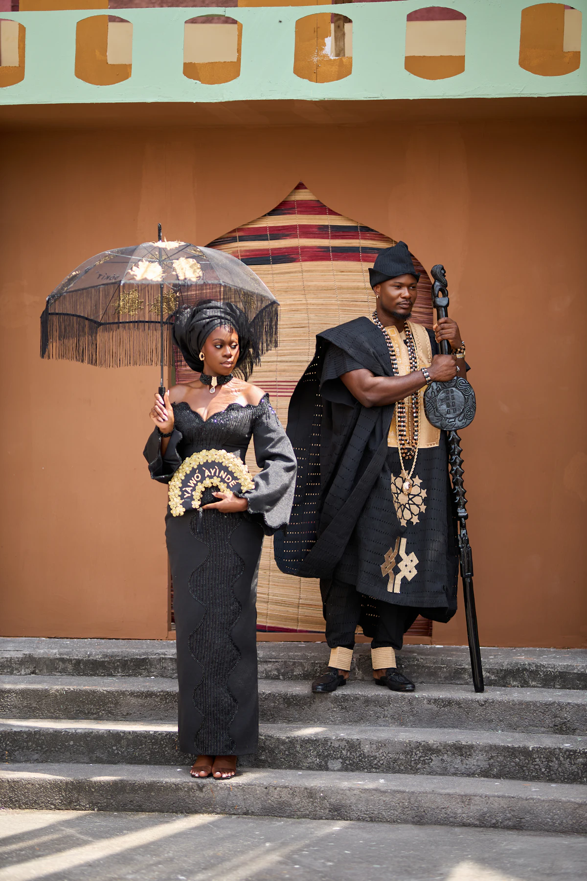 A couple in traditional attire, a woman with an umbrella and a man with a staff