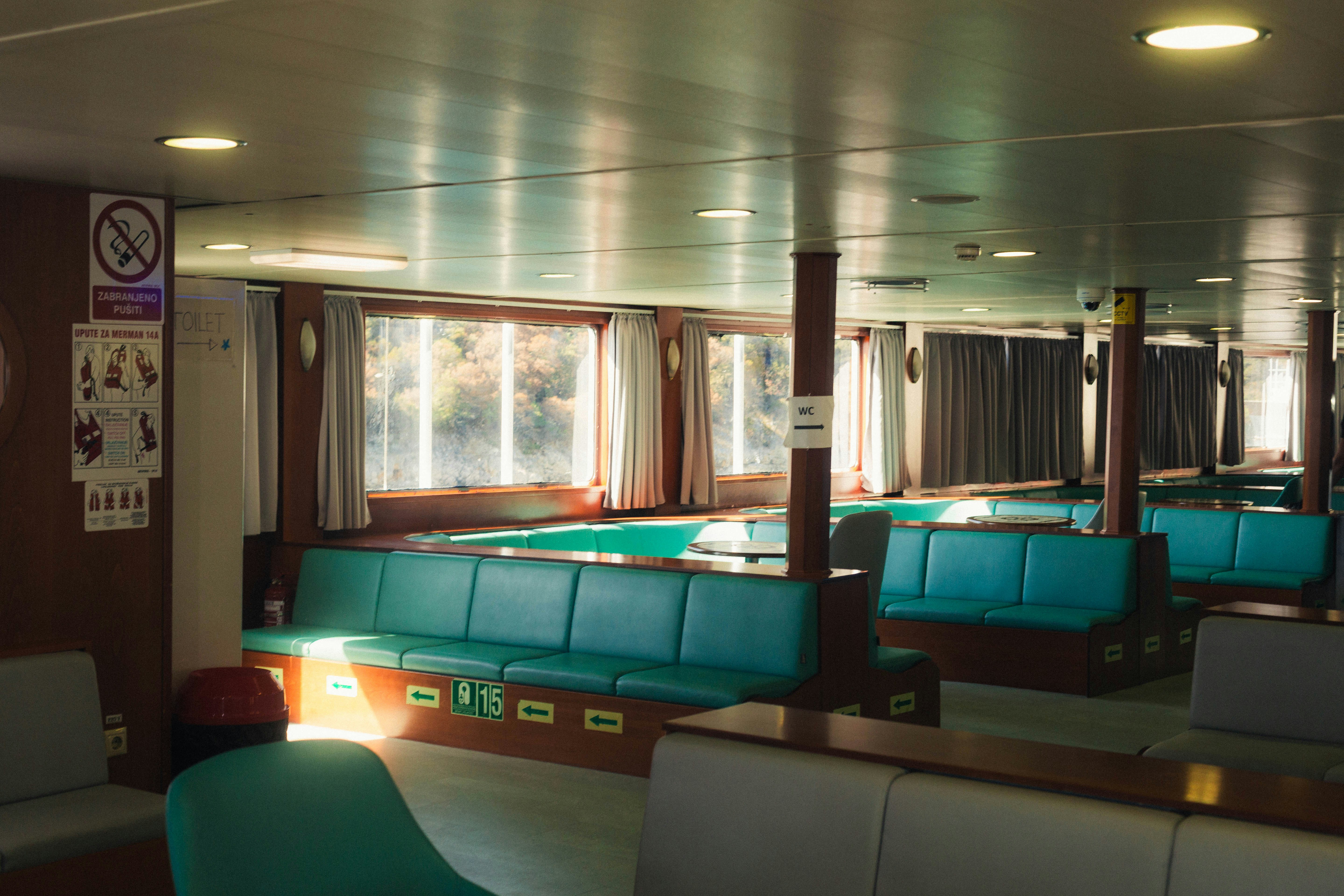 Empty turquoise bench seating inside a ferry cabin with large windows and curtains