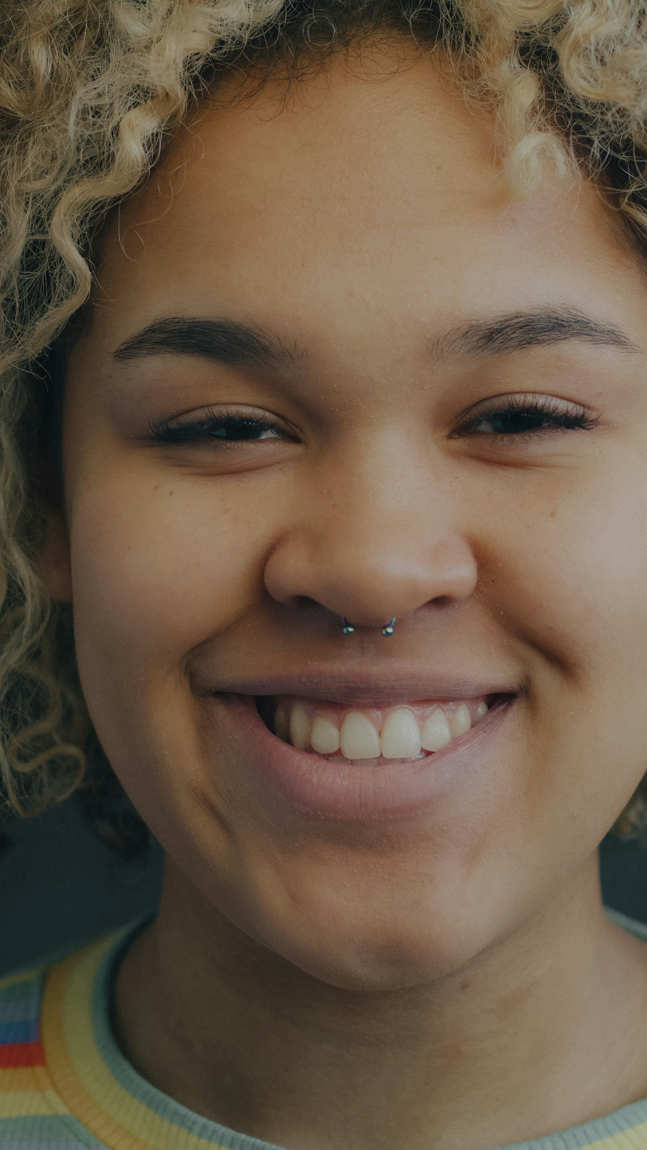 A smiling person with curly blonde hair and a silver septum piercing