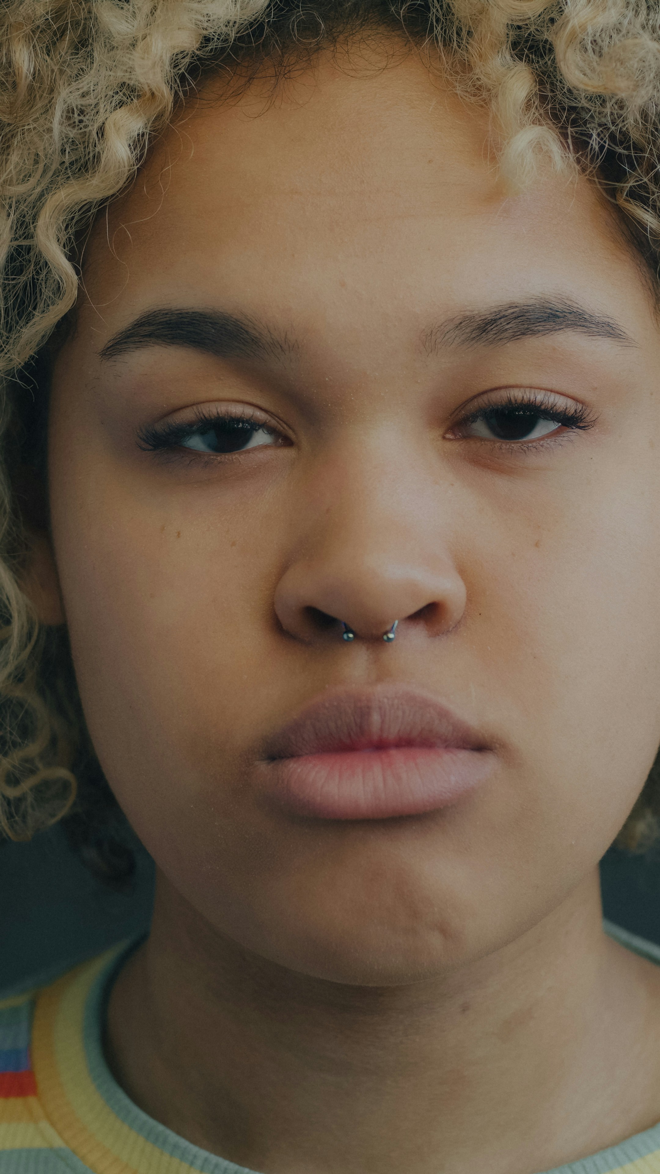 A person with curly blonde hair and a septum piercing looking forward