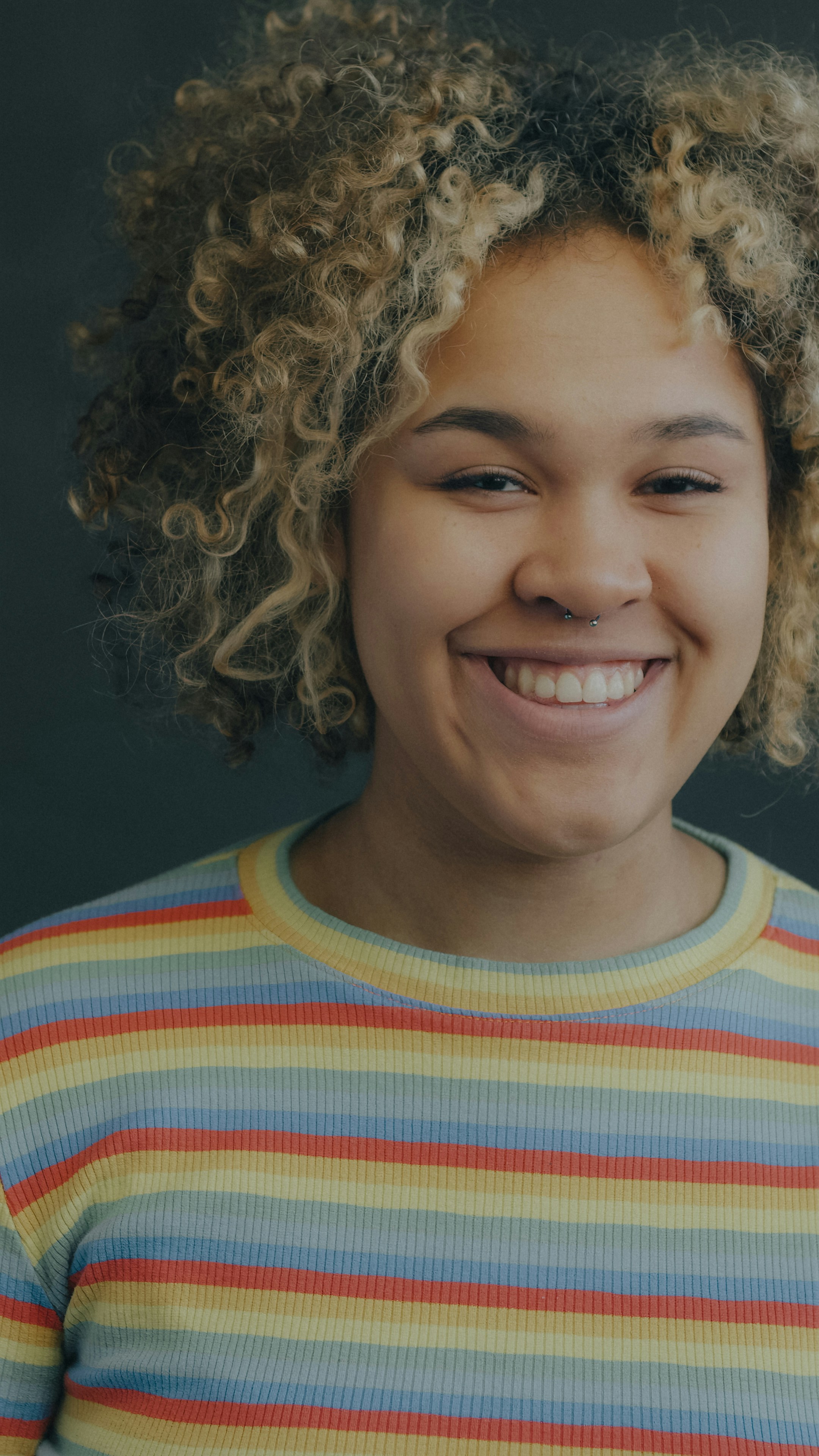 A smiling person with curly blonde hair and a septum piercing wearing a striped shirt