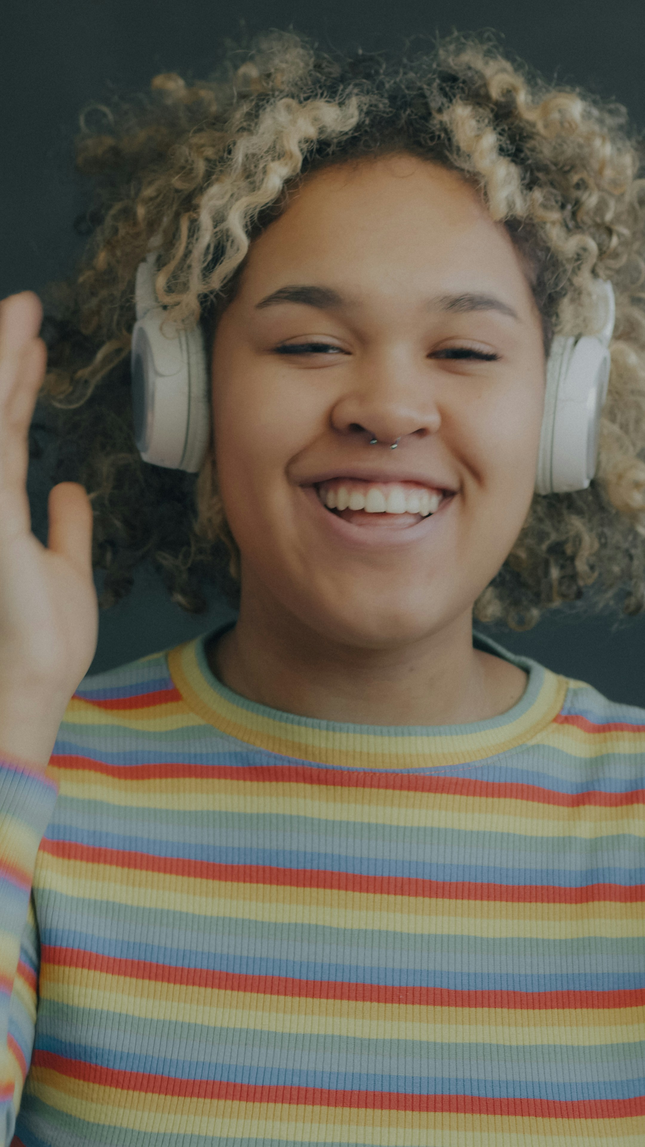 A person with curly blonde hair wearing white headphones waving at the camera