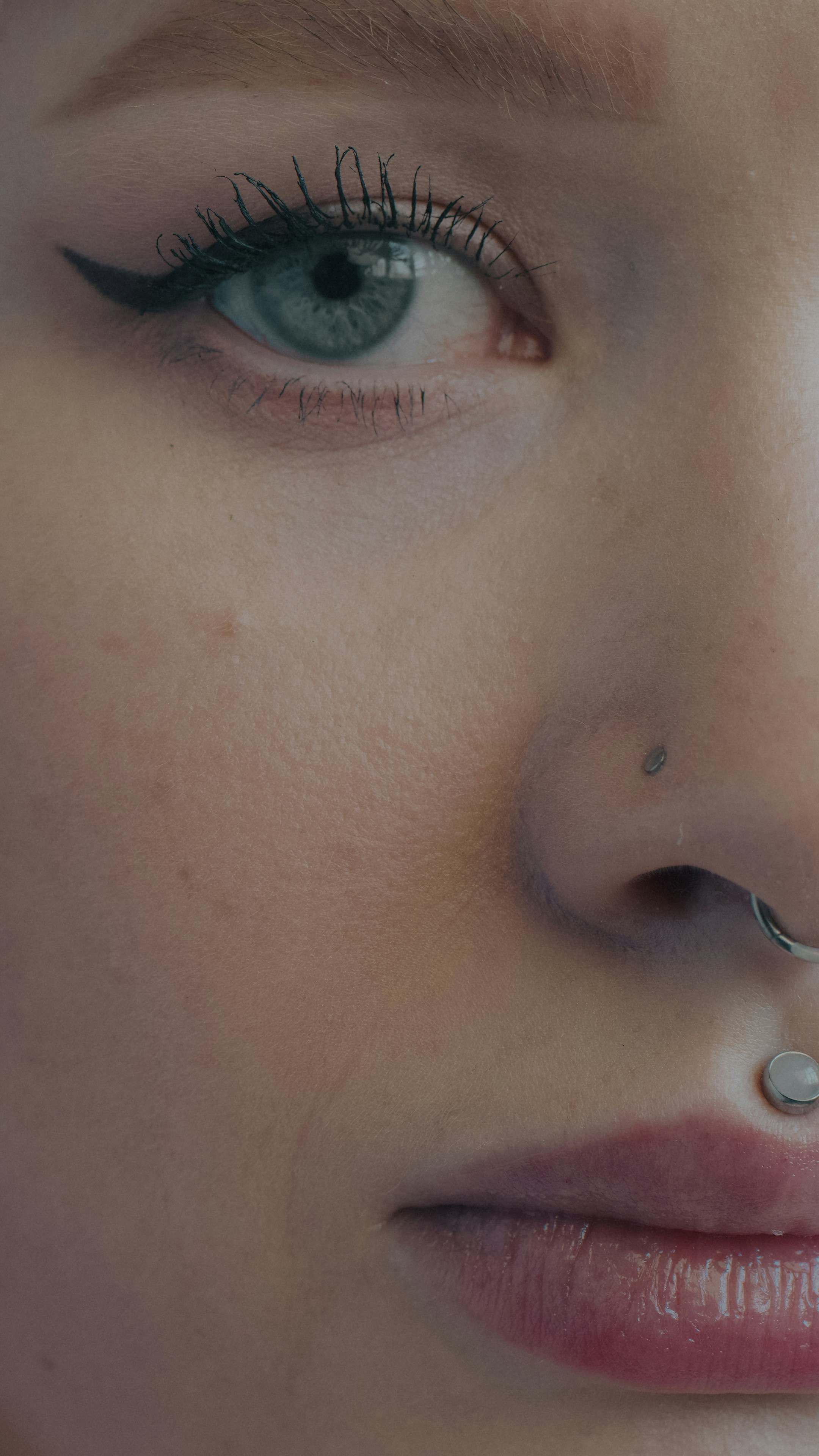 A close-up of a person with blue eyes, winged eyeliner, and facial piercings