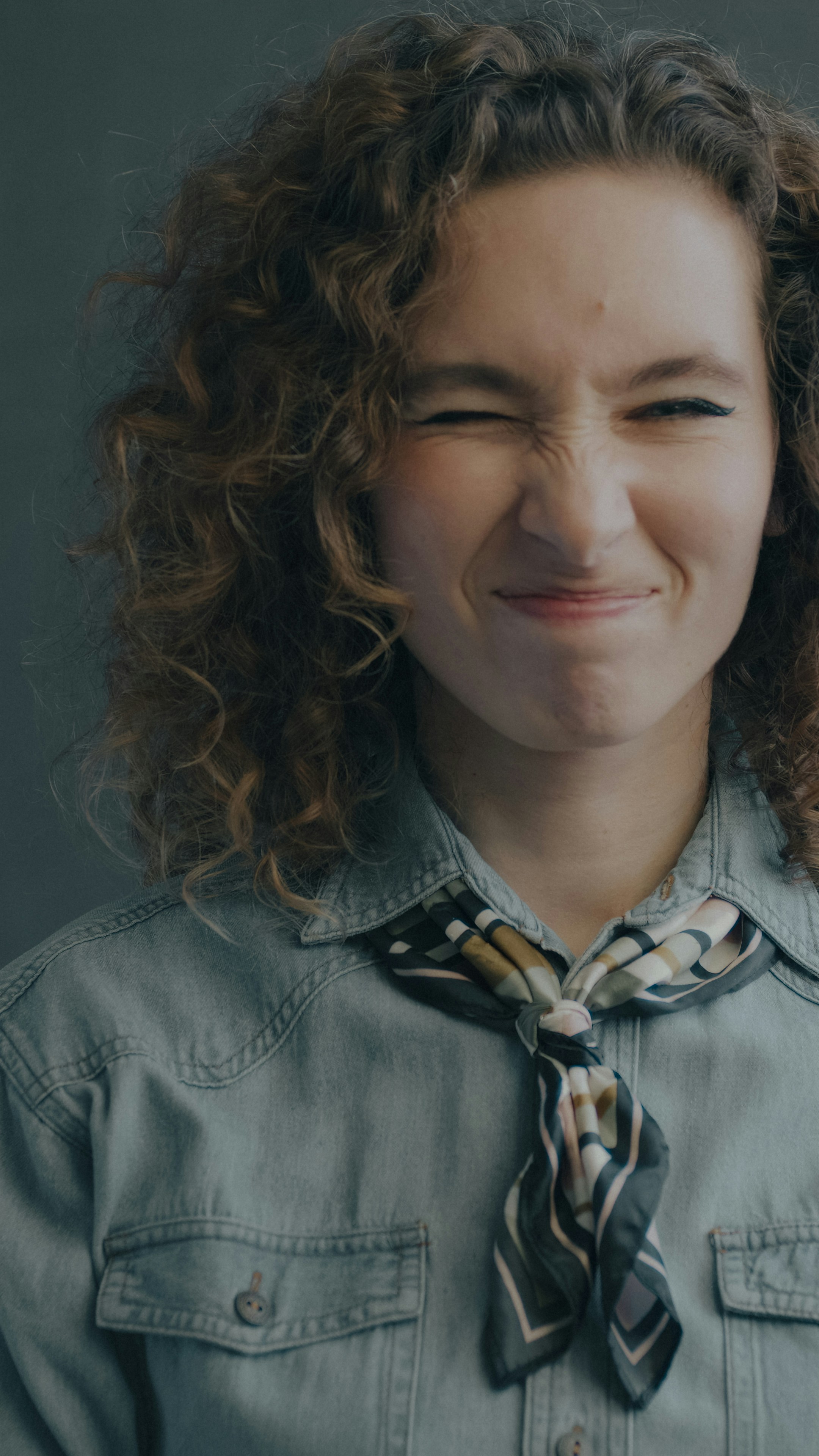 A woman with curly hair wearing a denim shirt and a patterned neckerchief