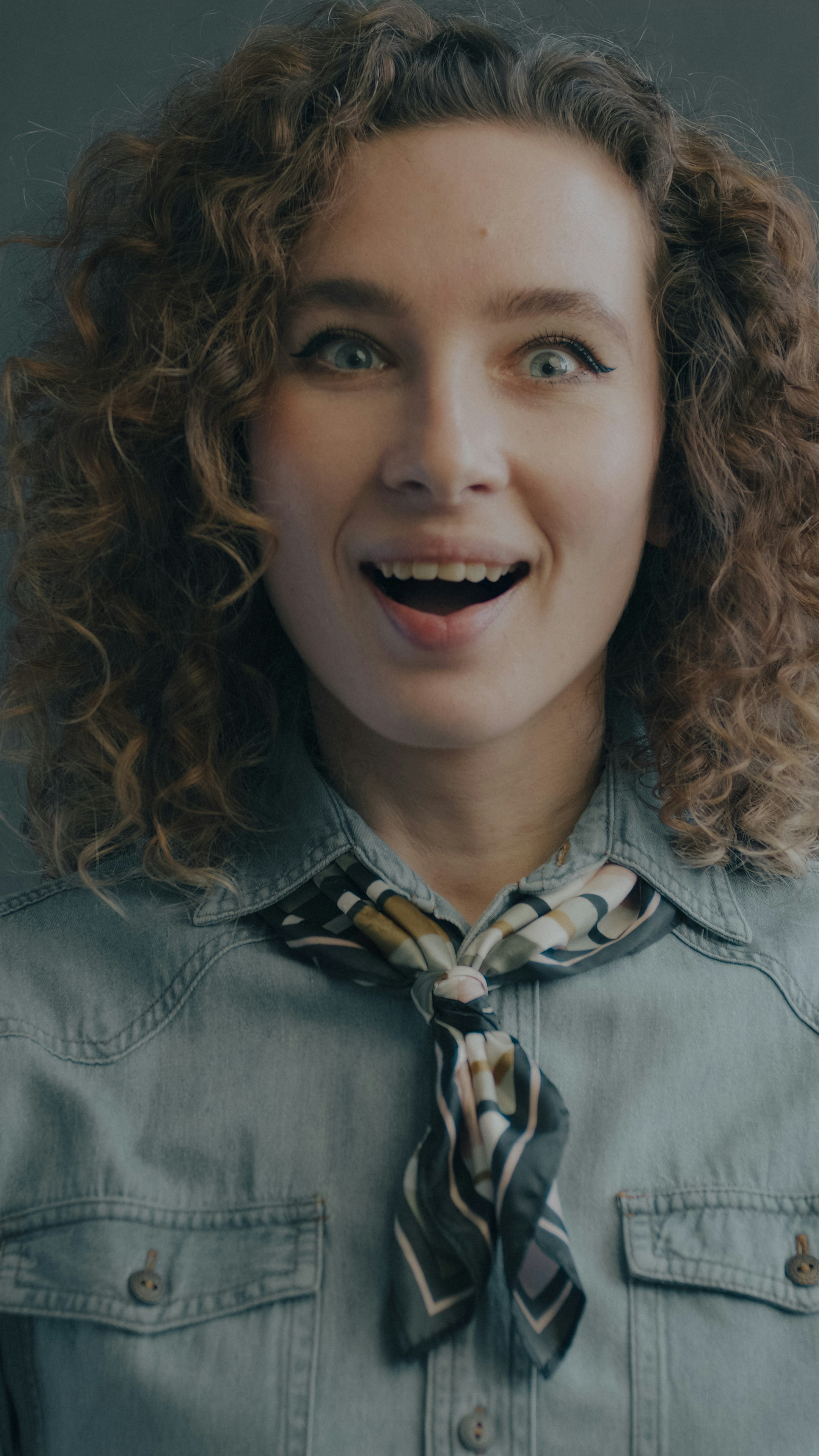 A woman with curly hair wearing a denim shirt and a patterned neck scarf