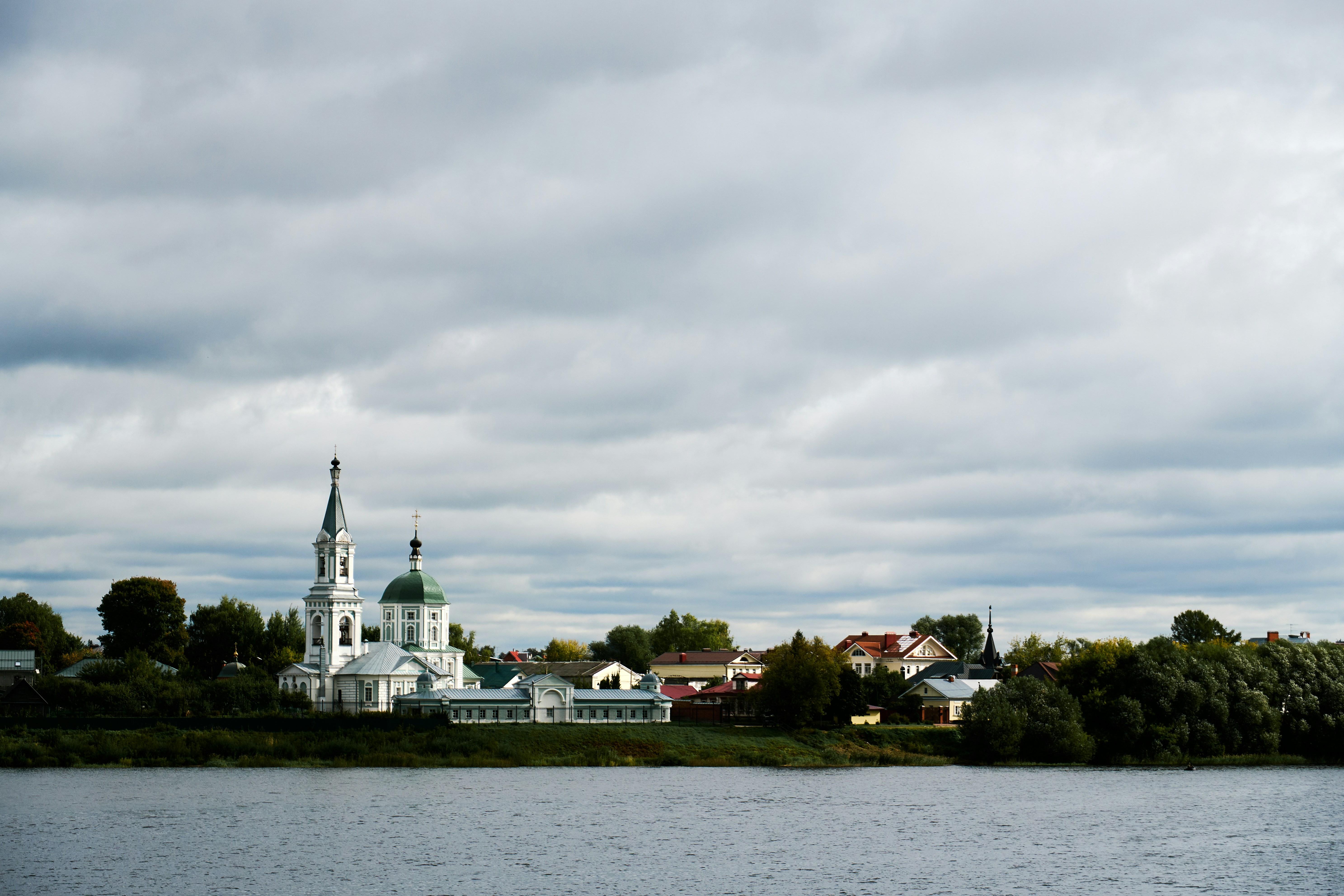 An Orthodox church with green domes on a riverbank under a cloudy sky