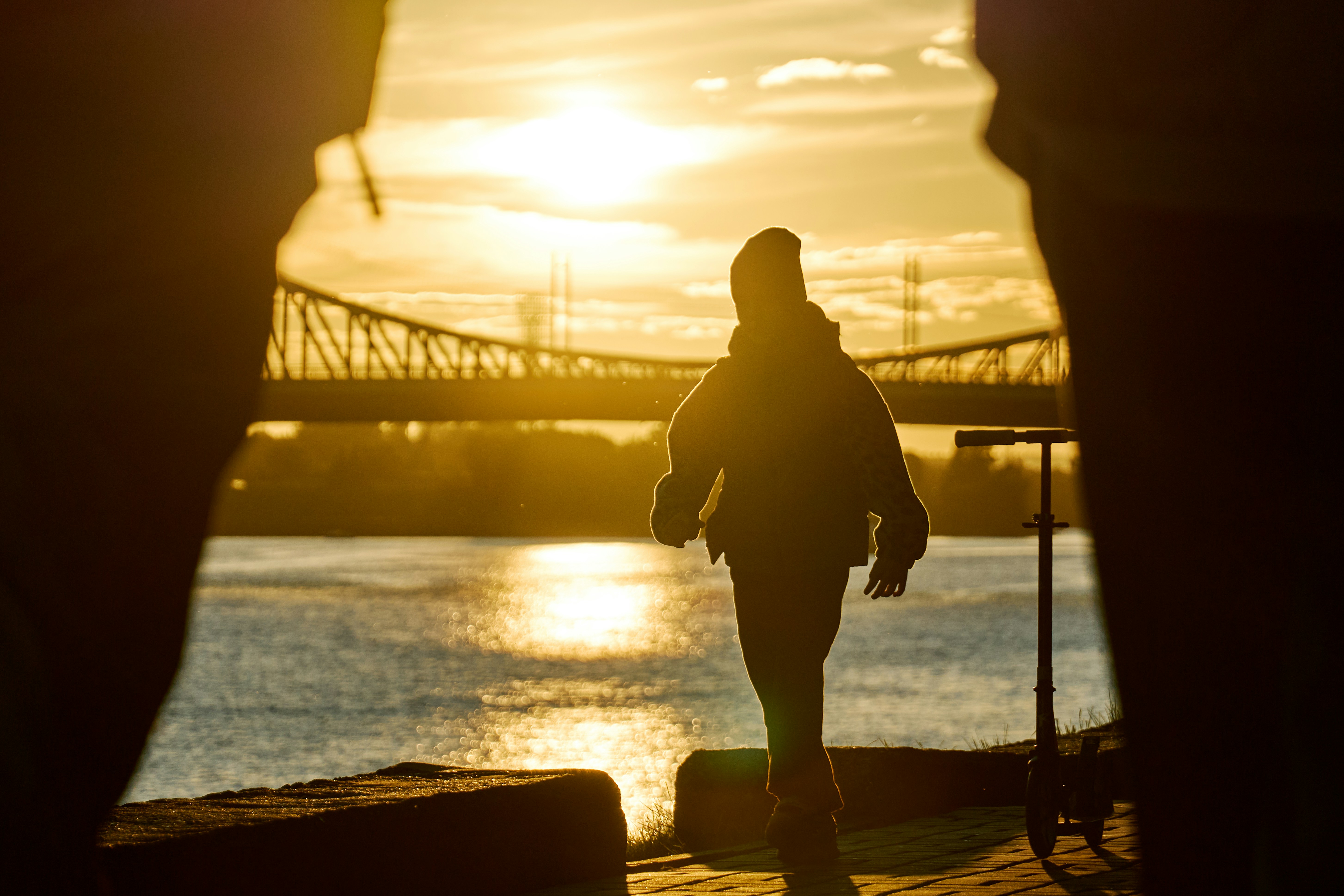 A person in silhouette with a scooter by a river at golden hour