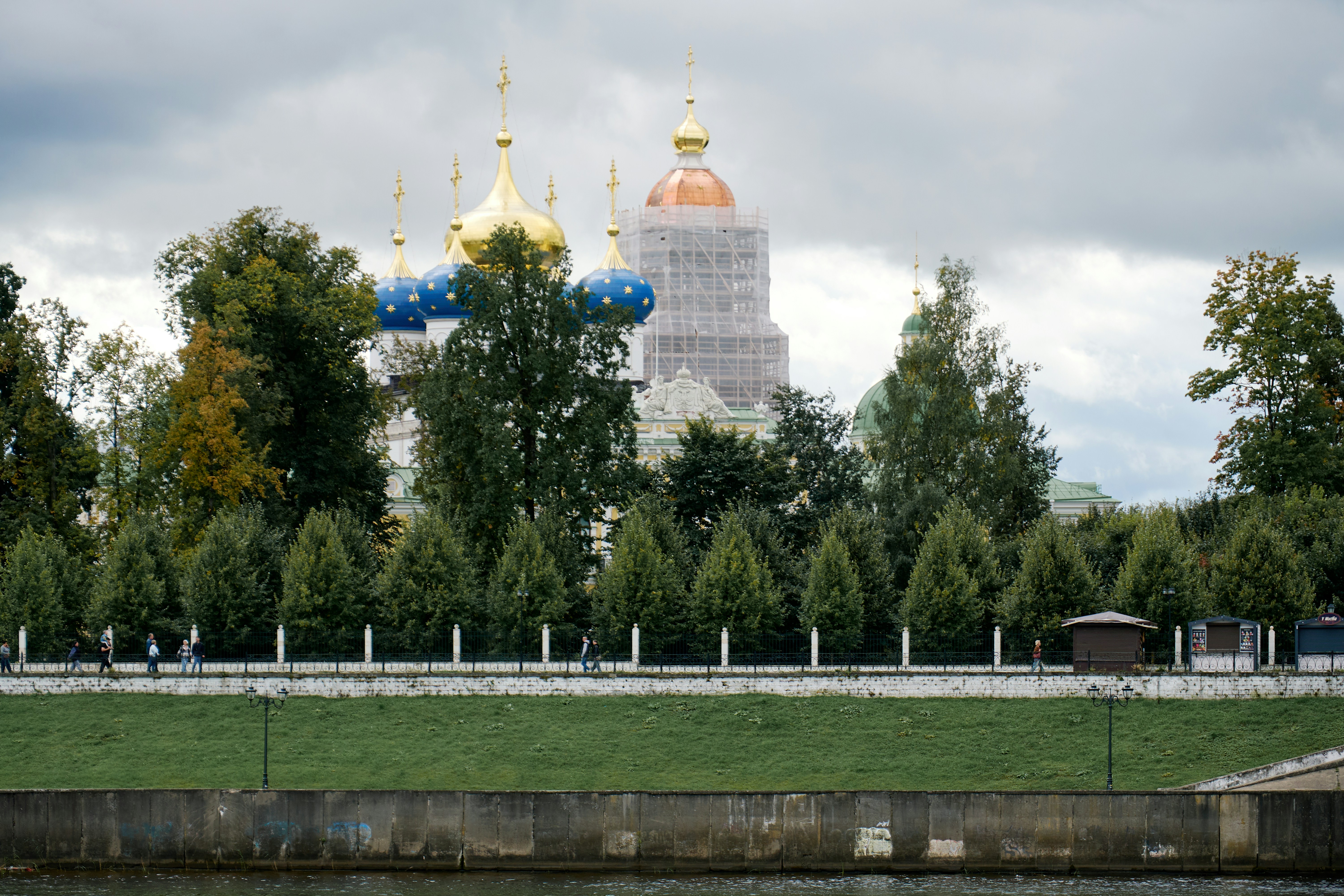 People walking along a path beside a river with an Orthodox cathedral behind trees