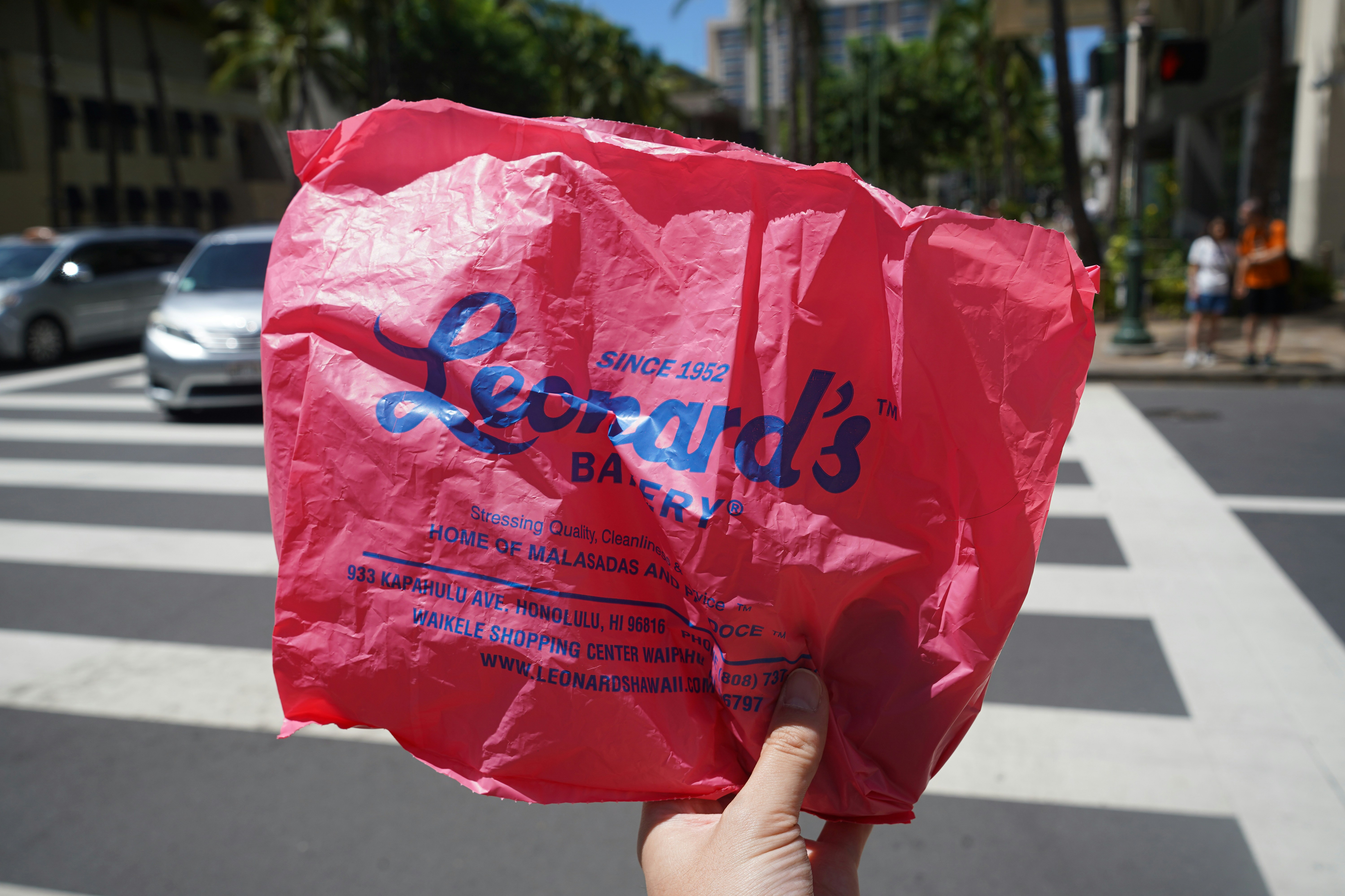 A hand holding a pink plastic bag from Leonard's Bakery at a crosswalk