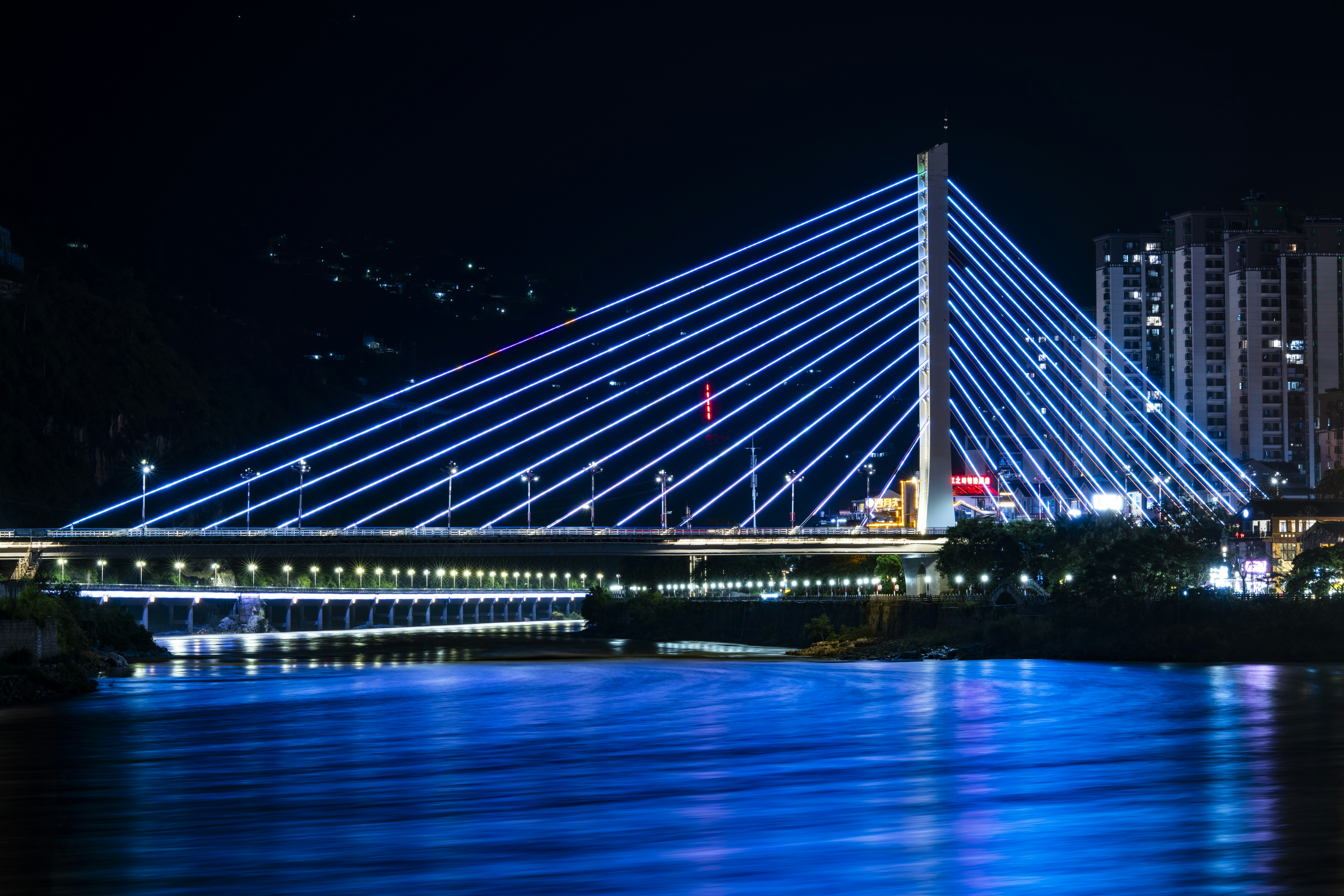 A cable-stayed bridge illuminated with blue neon lights over a dark river at night