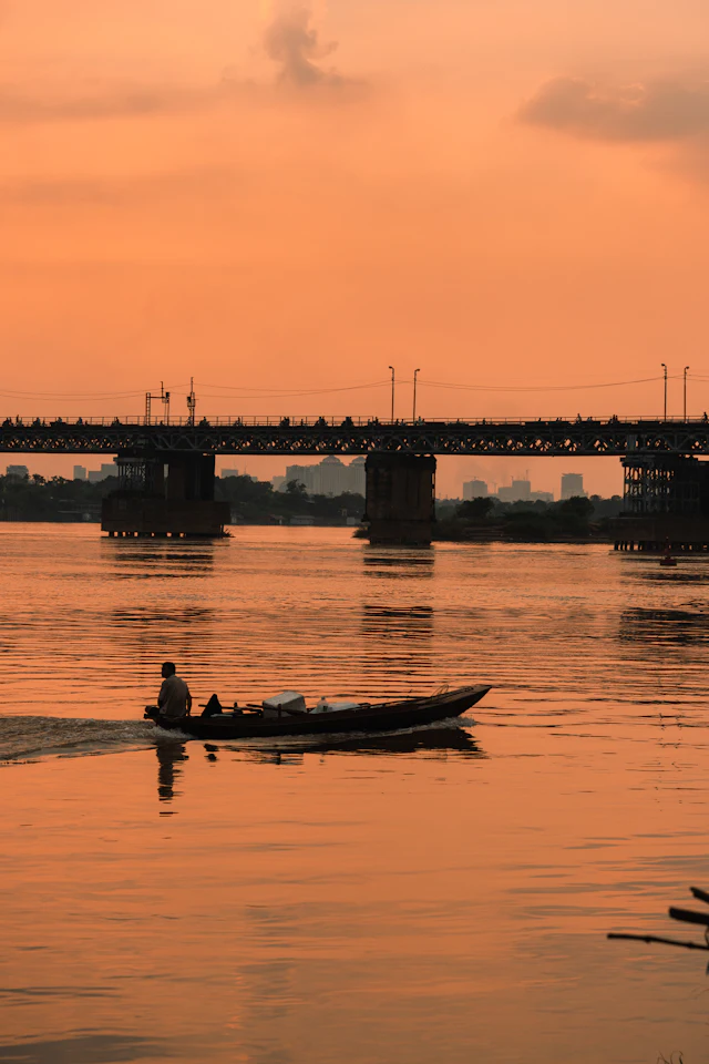 A small boat crosses the Red River beneath the silhouette of Long Bien Bridge under a deep orange sunset sky.