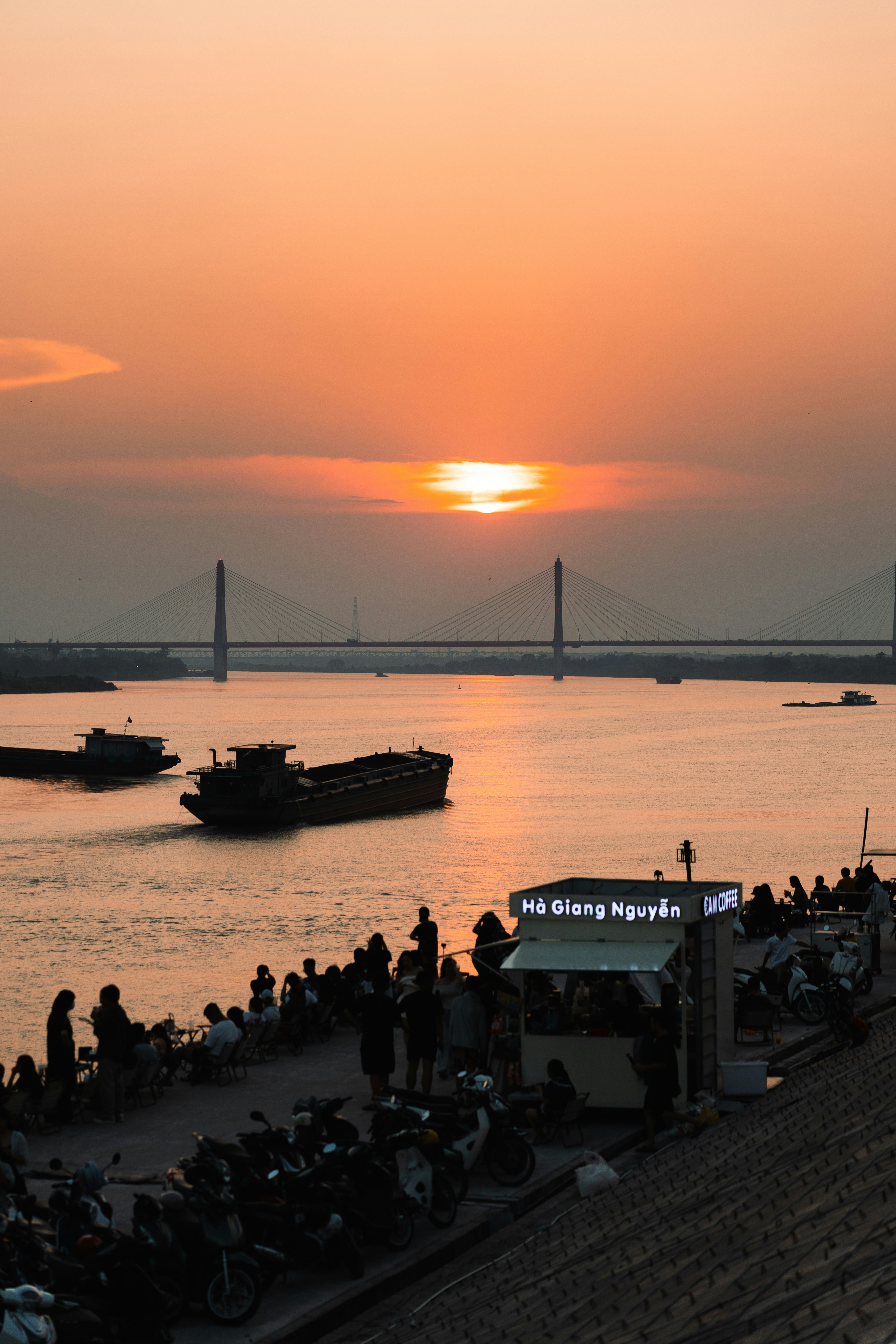 People gathered at a riverside cafe with a cable-stayed bridge under low sun