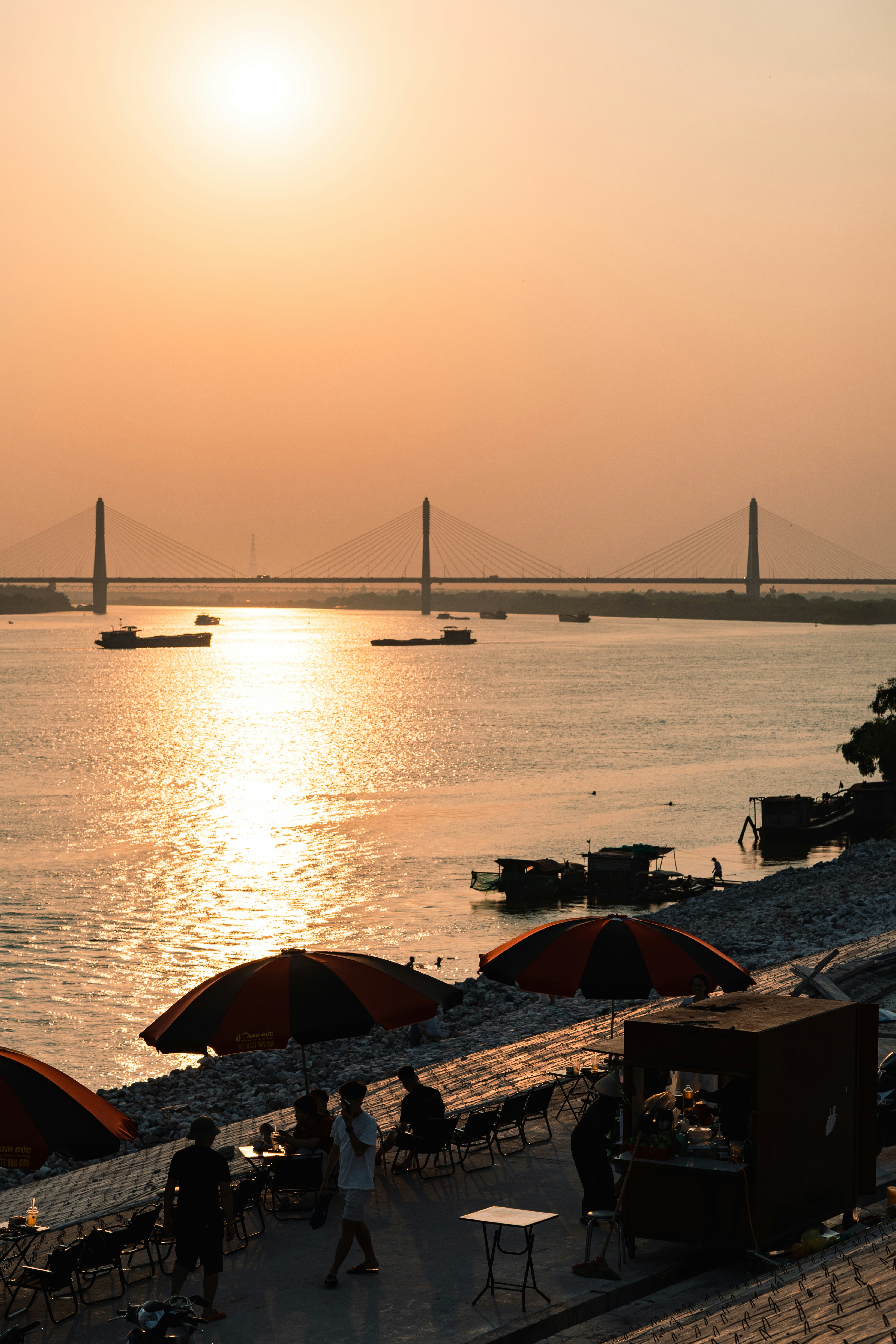 People sitting under umbrellas by a river with a cable-stayed bridge at low sun