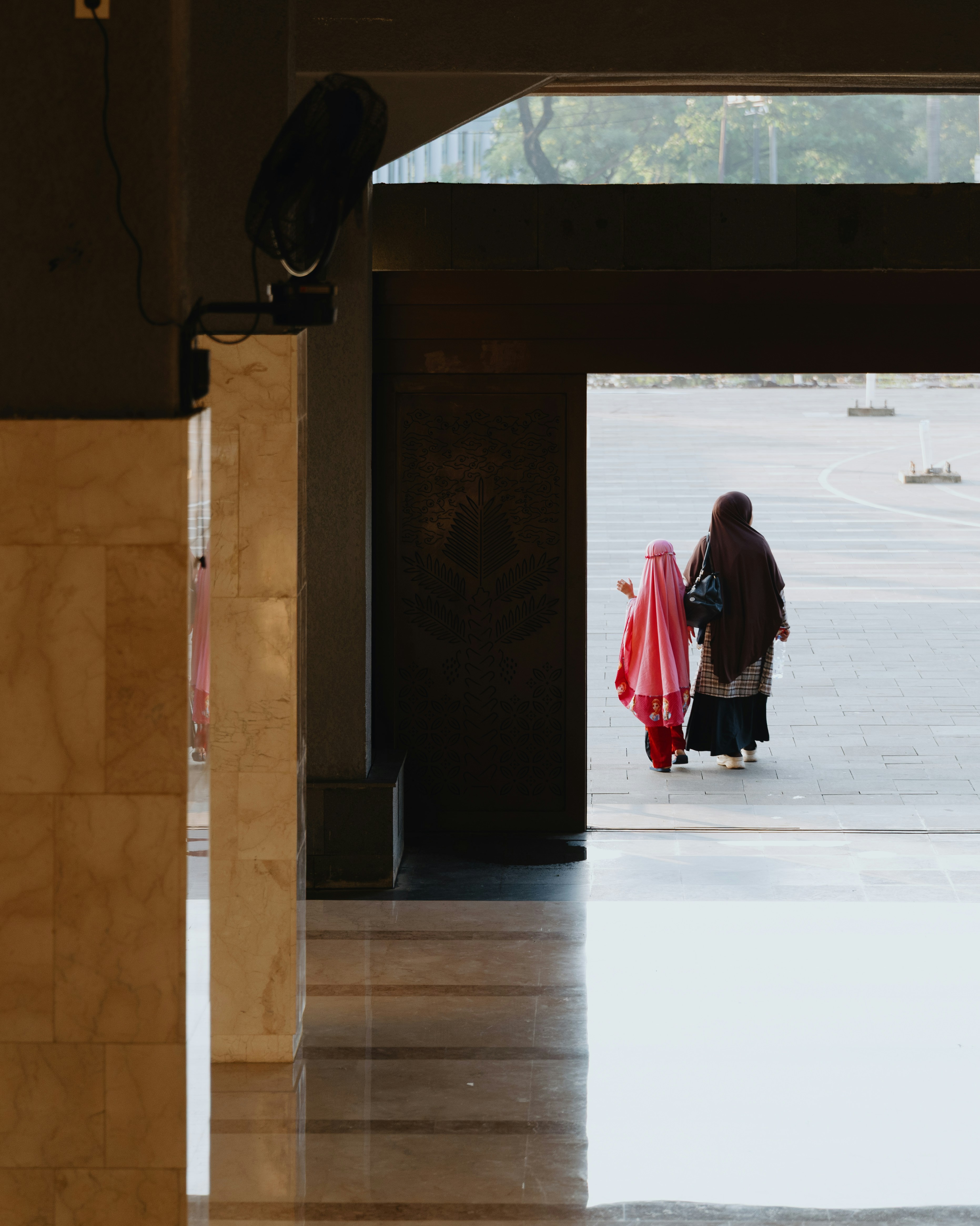 An adult and a child in headscarves walking through a bright doorway in Bogor