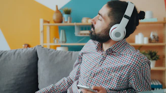 A man wearing white headphones in a plaid shirt on a grey sofa
