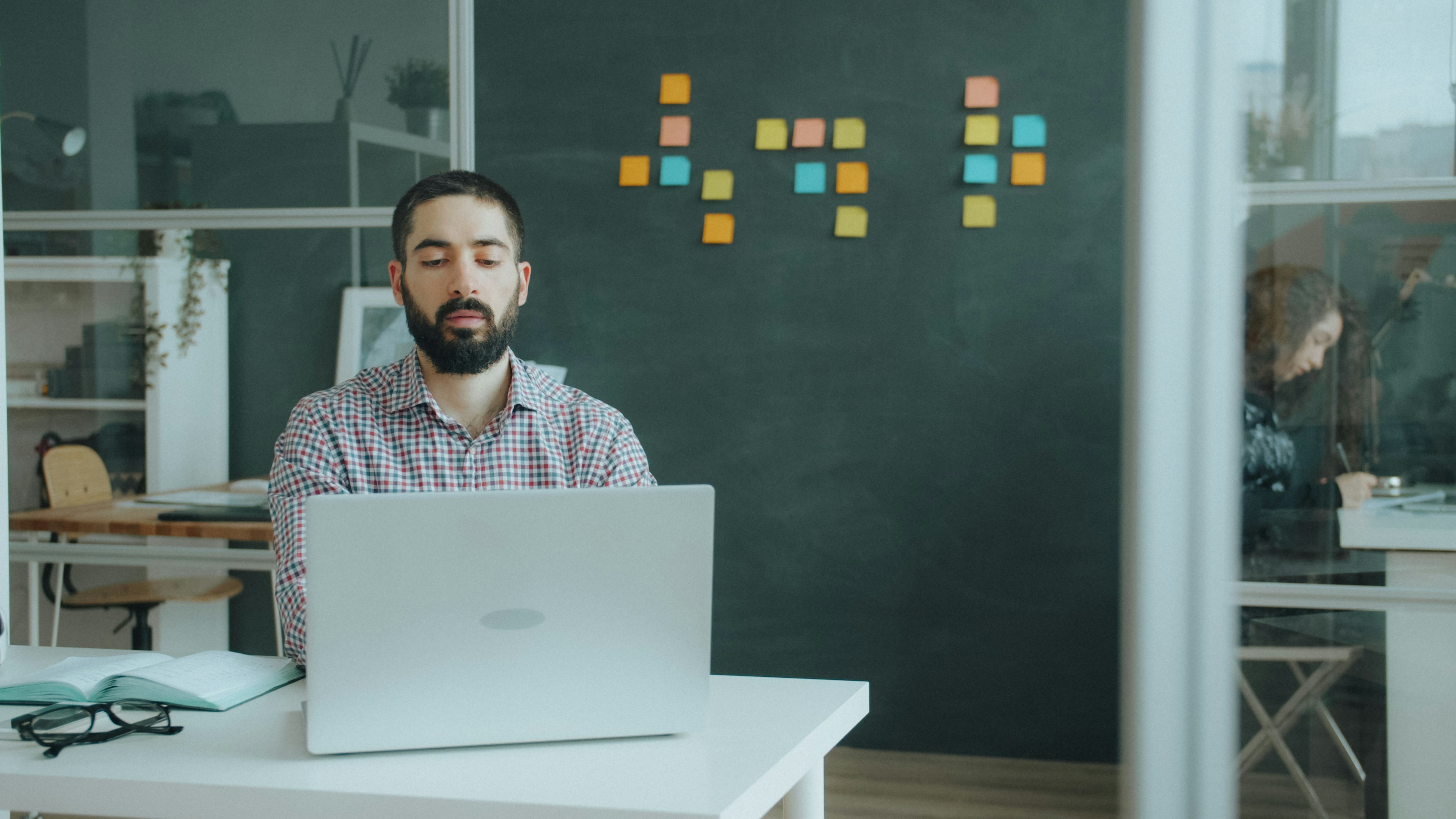 A man with a beard using a laptop at a desk in a workspace