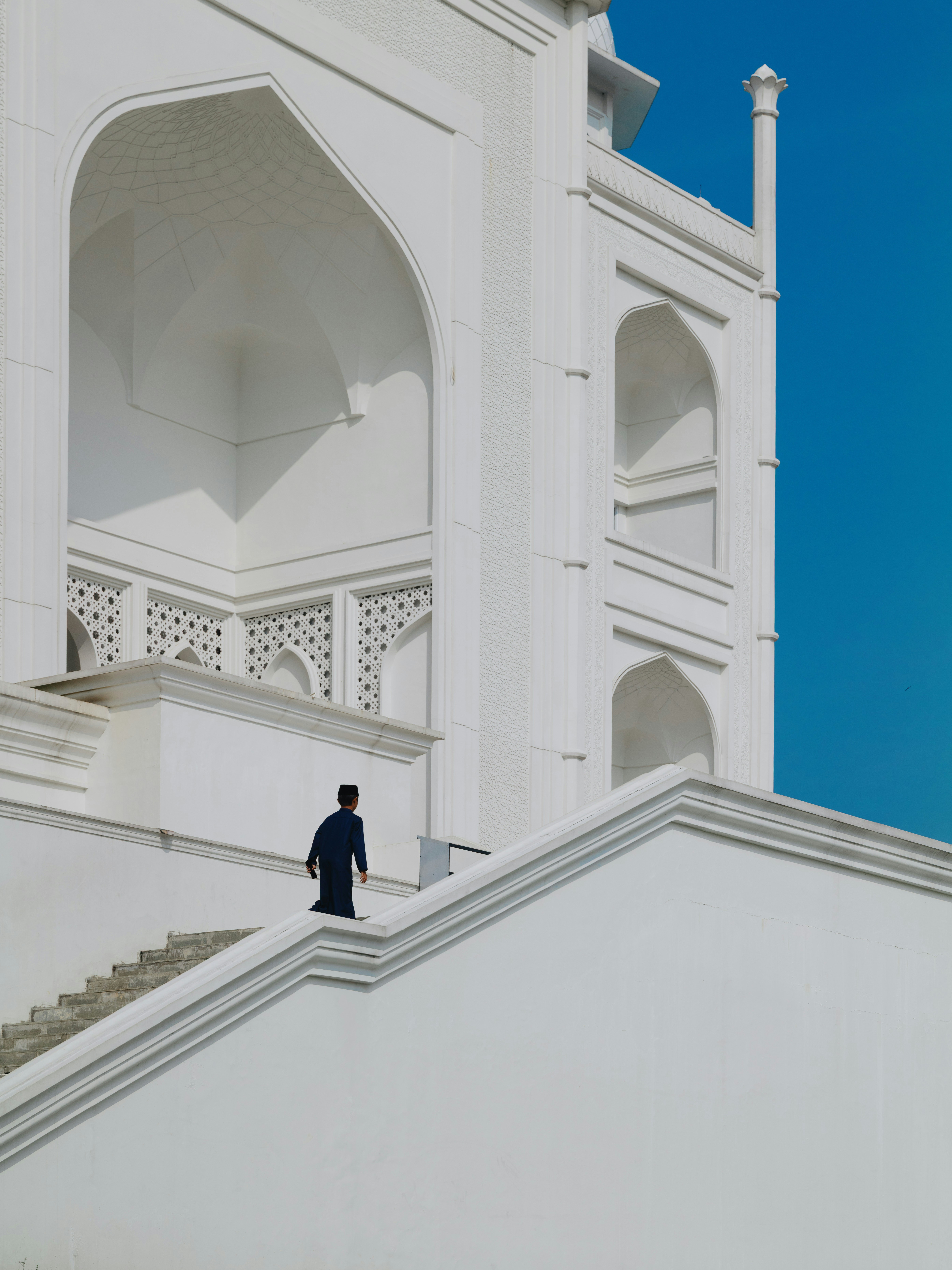 A person in a dark outfit walking up white stairs at Masjid Budi Guna