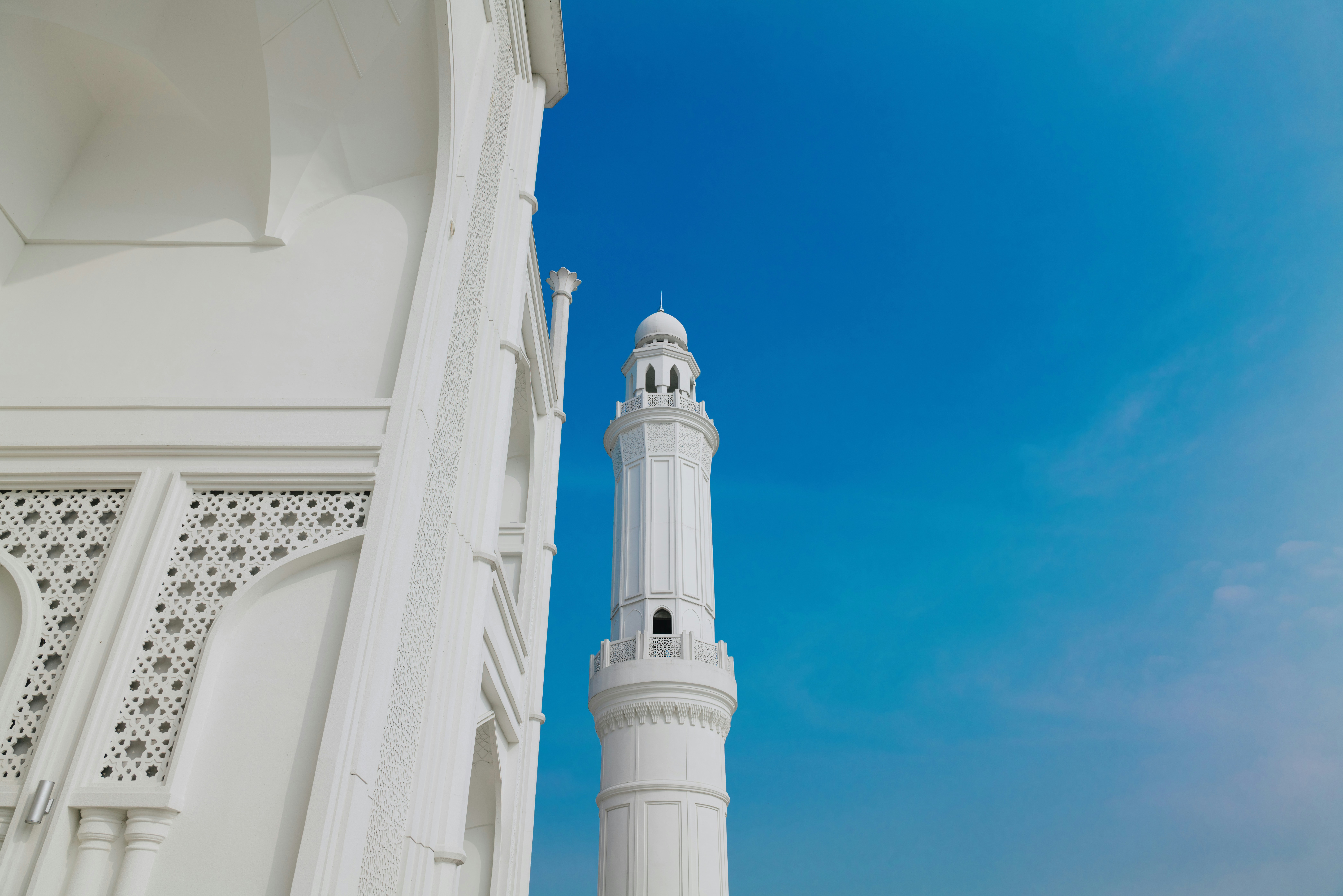 A white minaret beside the carved facade of Masjid Budi Guna against blue sky