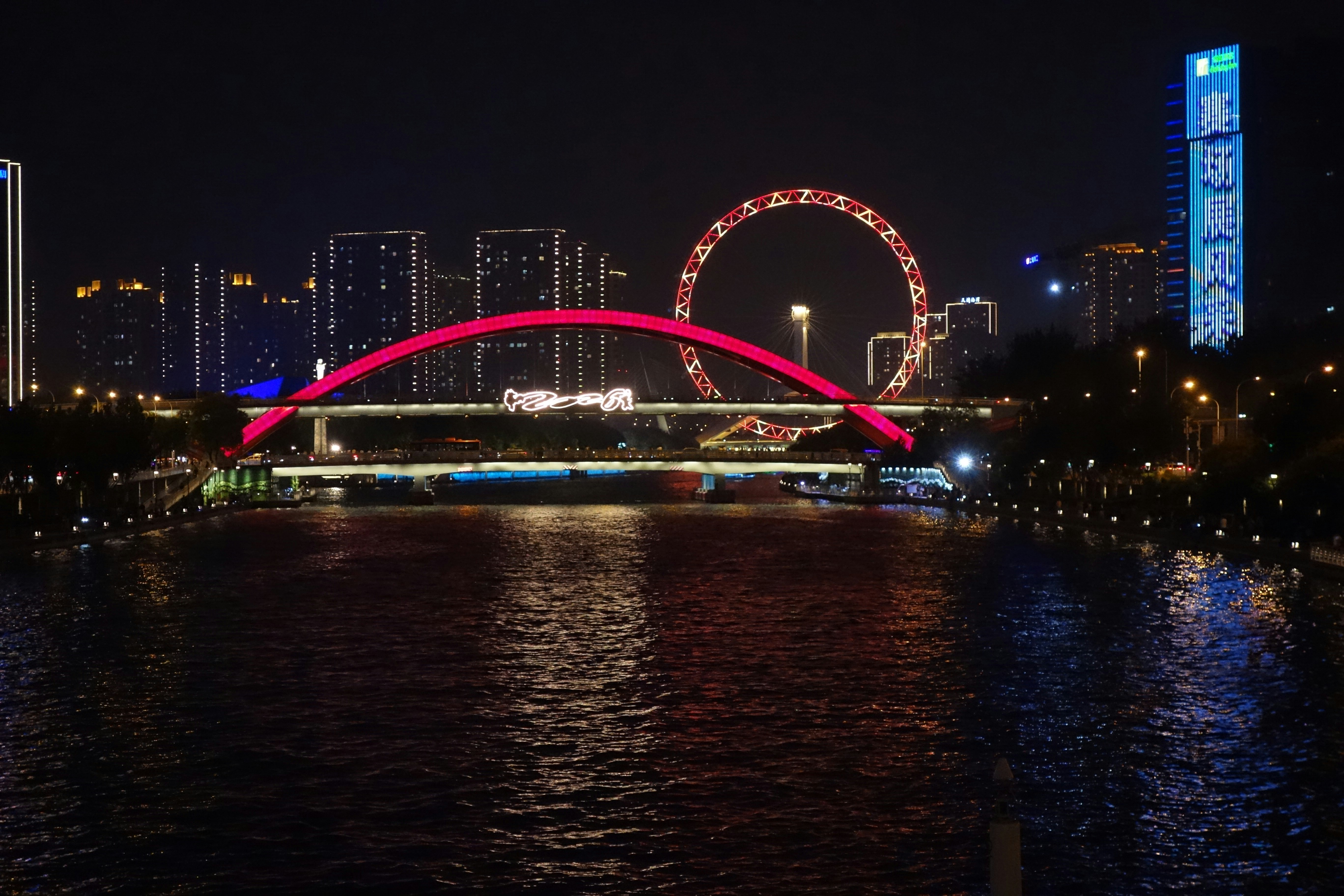 A red illuminated bridge and ferris wheel over a dark river at night