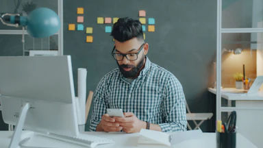 A man with a beard and glasses using a smartphone at a desk