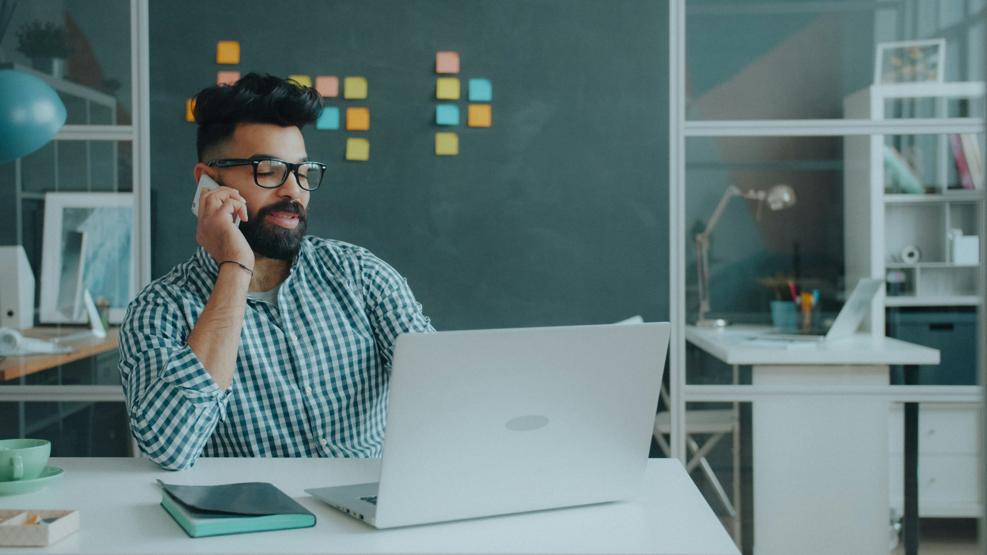 A man with a beard and glasses talking on a phone at a desk