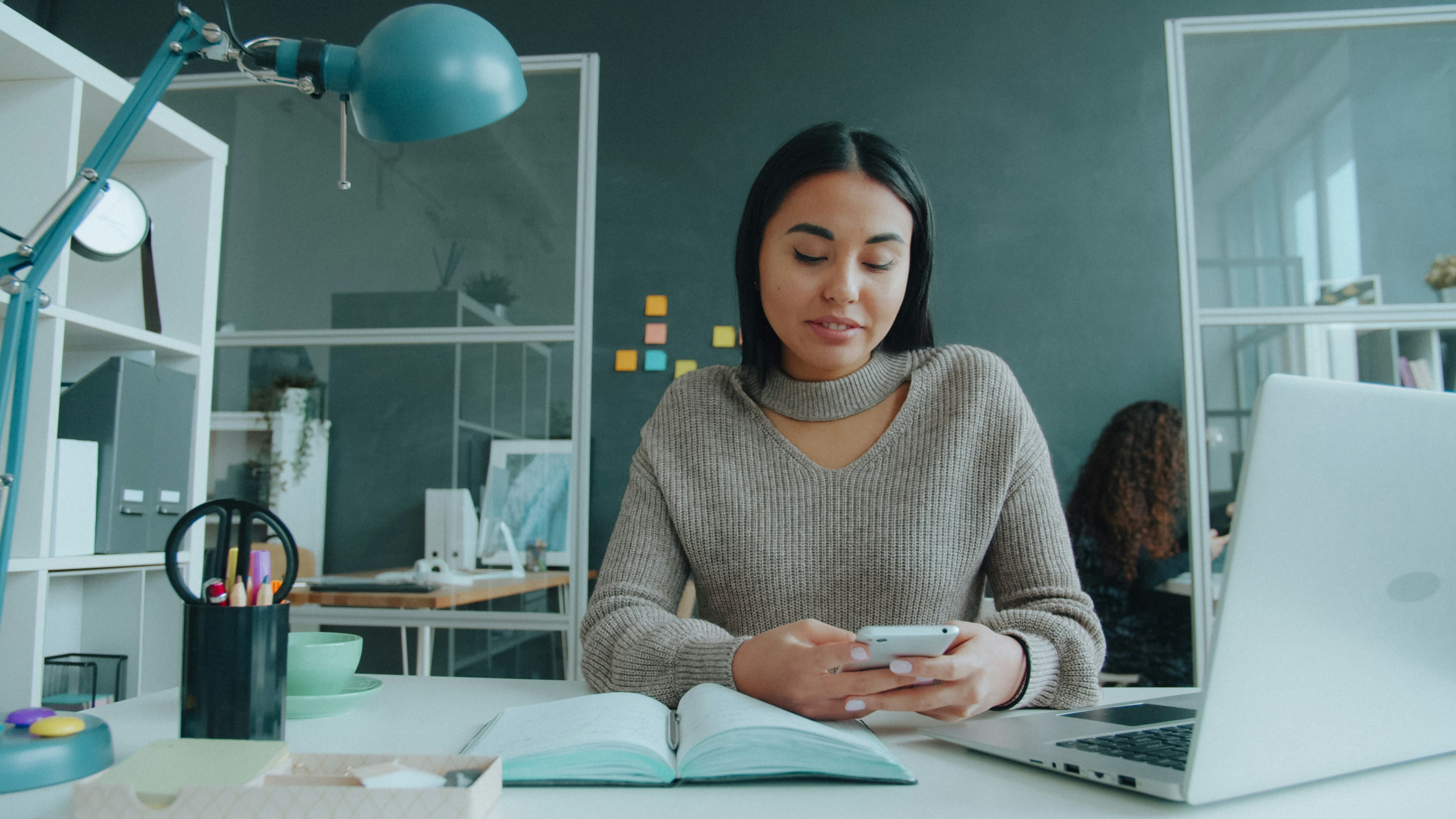 Portrait of beautiful mixed race lady texting touching smartphone screen in modern office while young woman is working in background. People and gadgets concept.