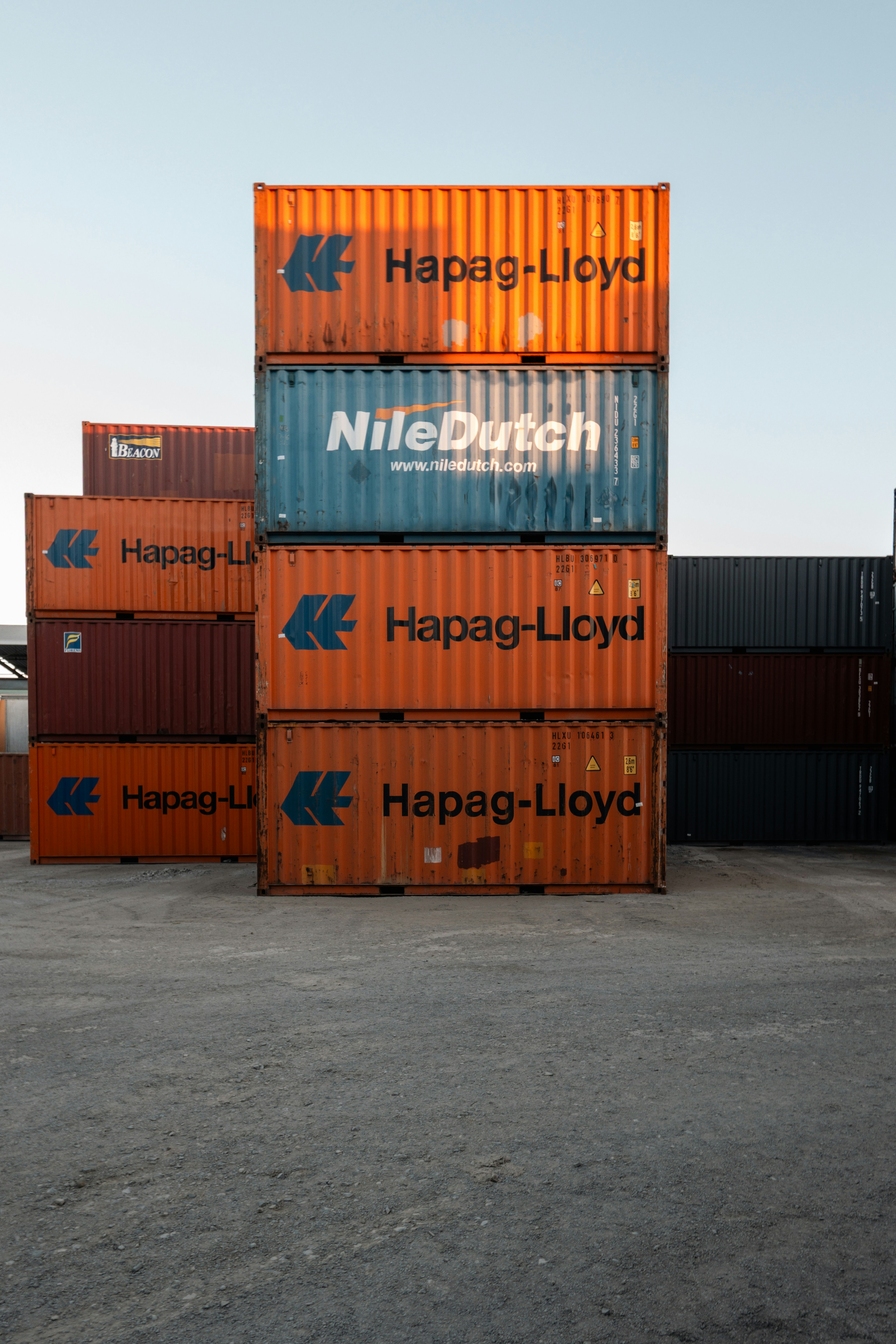 A vertical stack of orange Hapag-Lloyd shipping containers dominates a harbor lot, with blue Niledutch containers interleaved and a clear sky above. The scene emphasizes industrial scale and repetitive geometry.