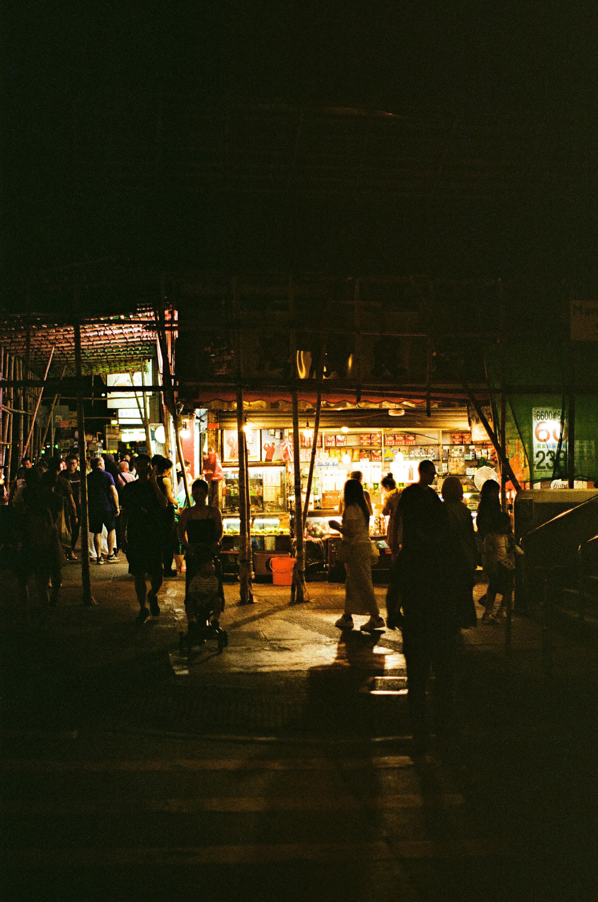 People walking past a brightly lit street stall at night under bamboo scaffolding