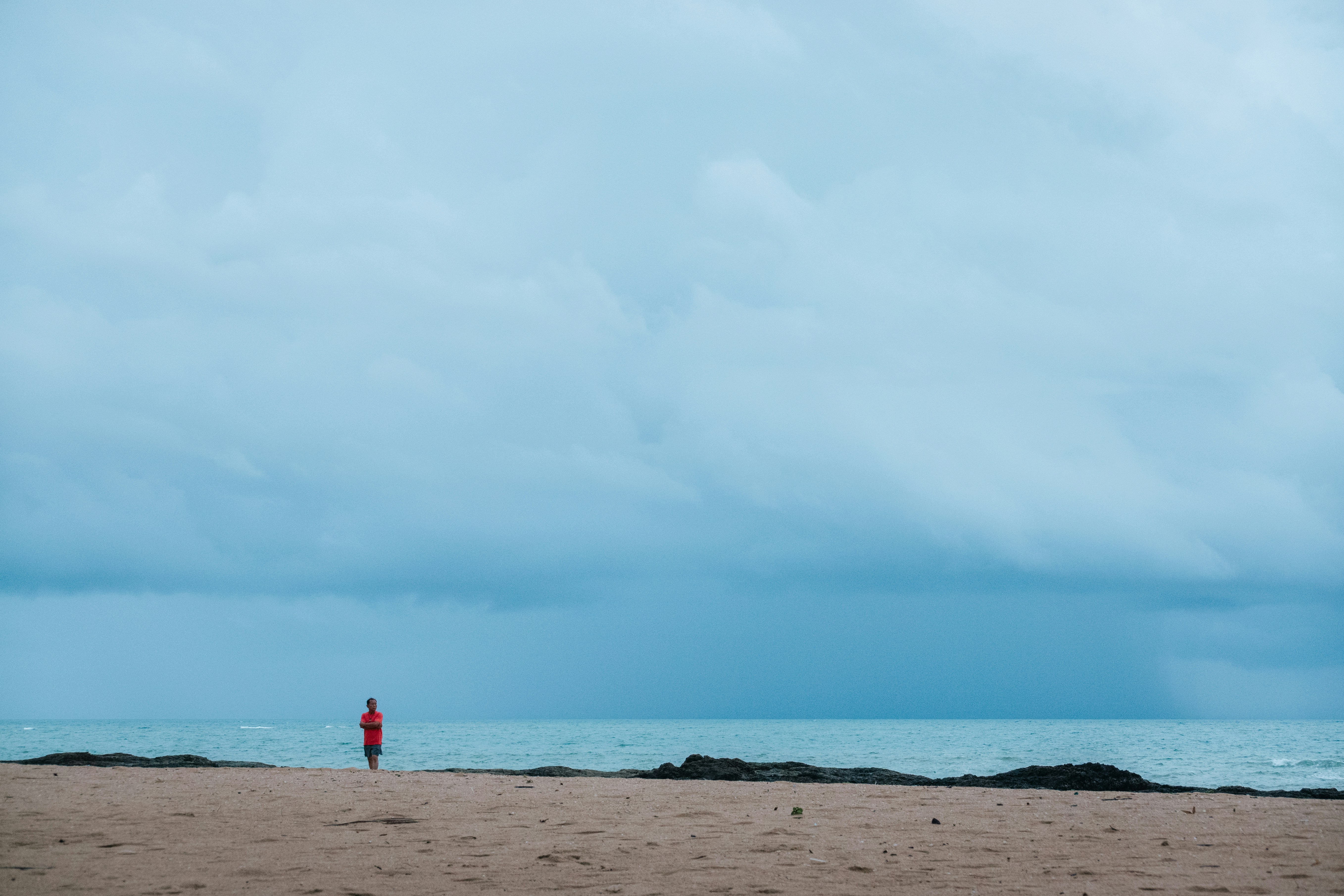 A person in a red shirt standing on a sandy beach under stormy clouds
