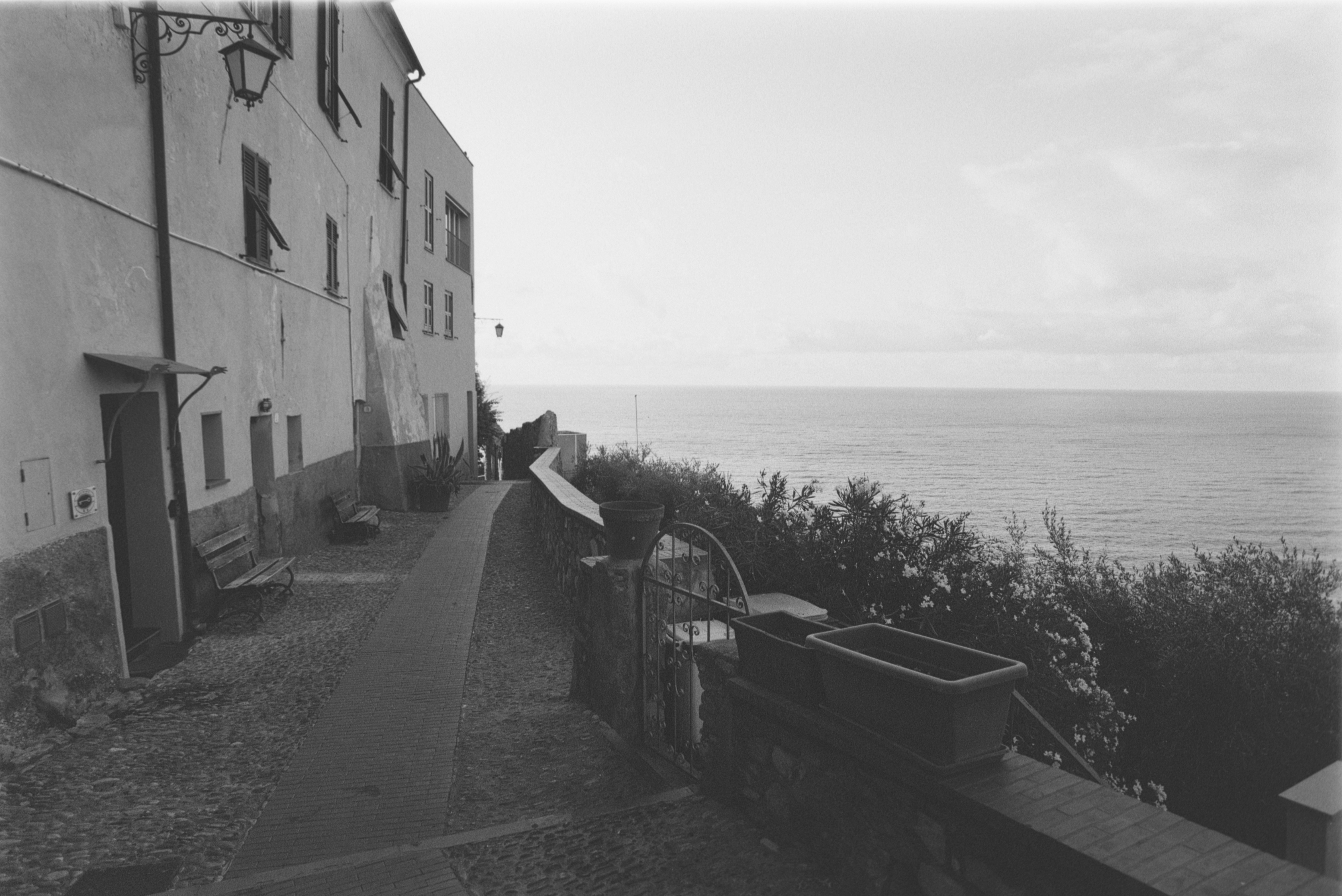 Ein steinerner Weg entlang einer Gebäudemauer mit Blick auf das Meer in Schwarz-Weiß