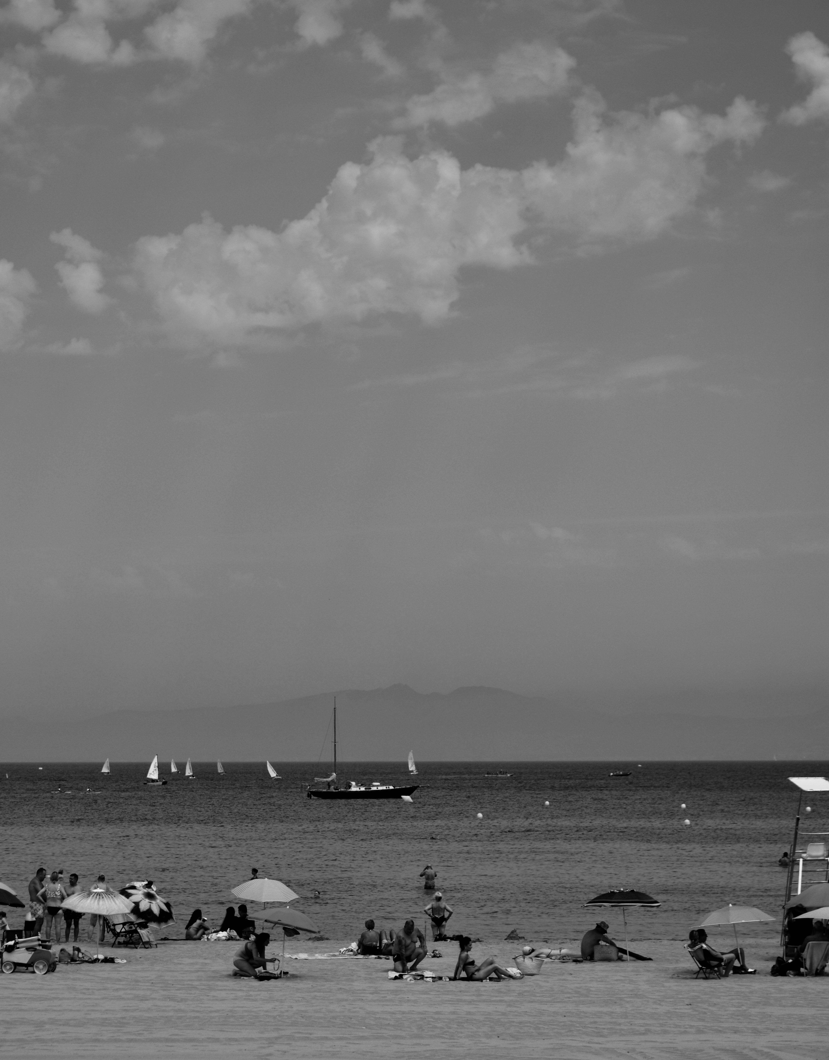 People on a sandy beach with umbrellas and sailboats on the water
