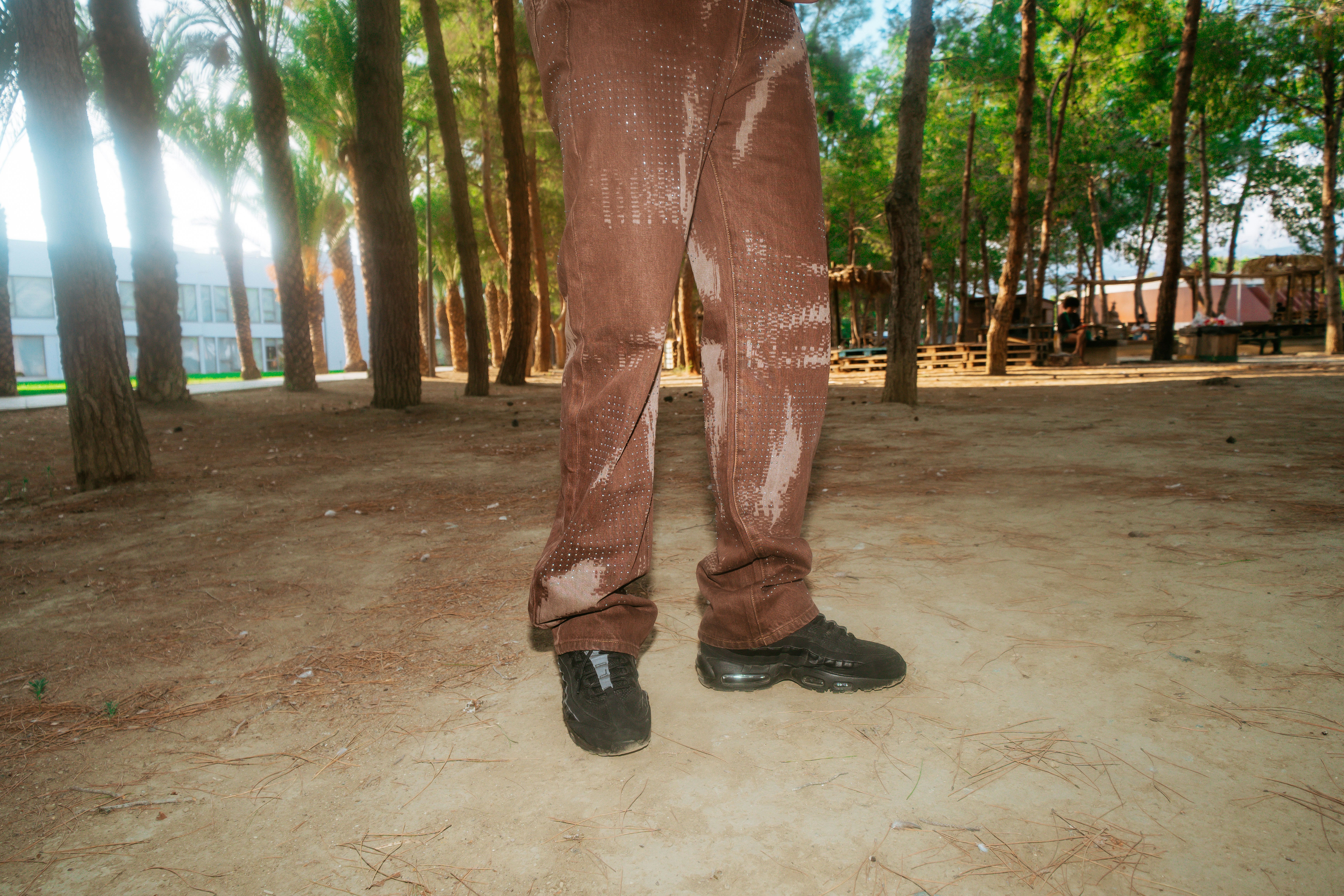 A person wearing brown patterned jeans and black sneakers in a pine forest