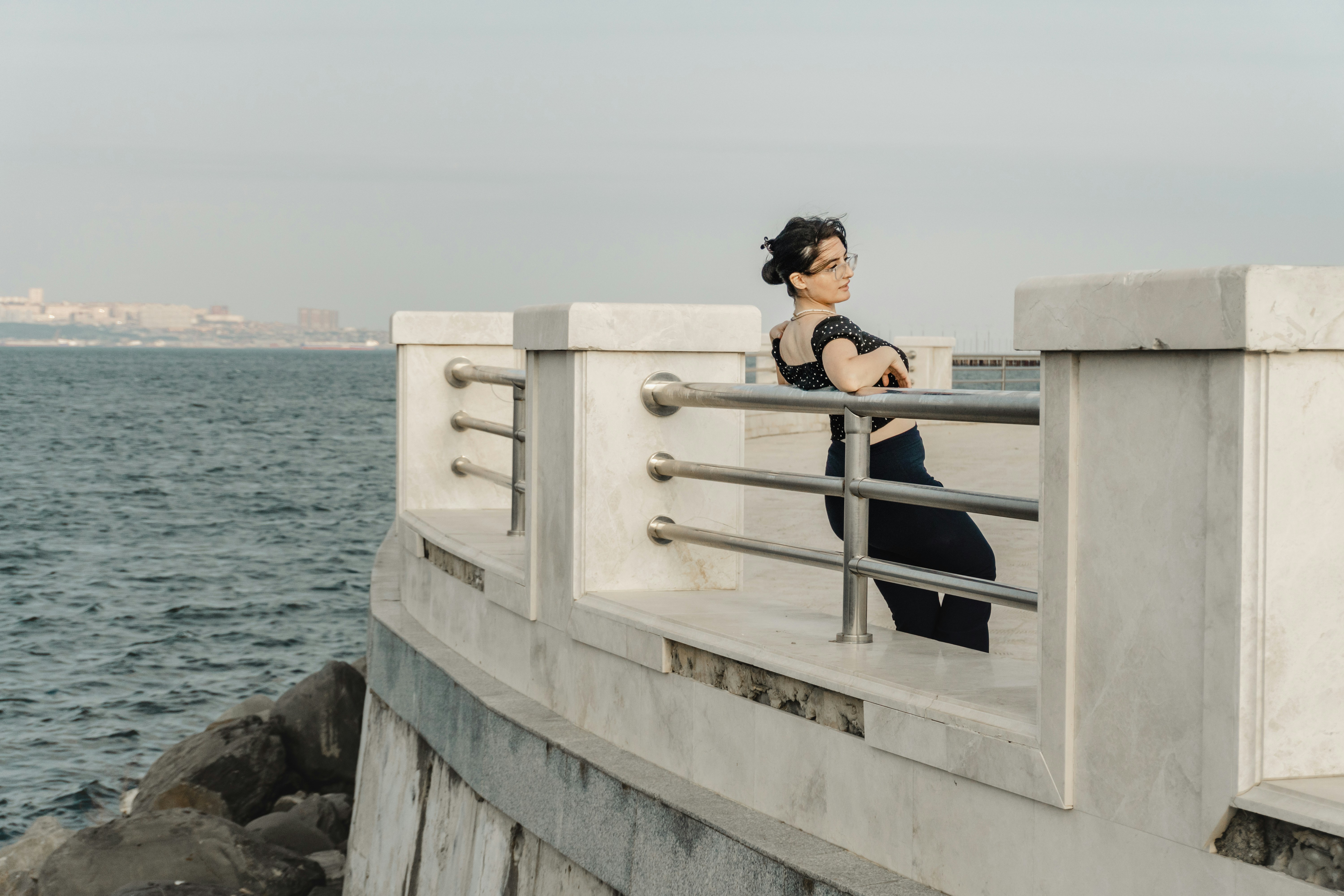 A woman in glasses leaning against a stone railing along a waterfront promenade